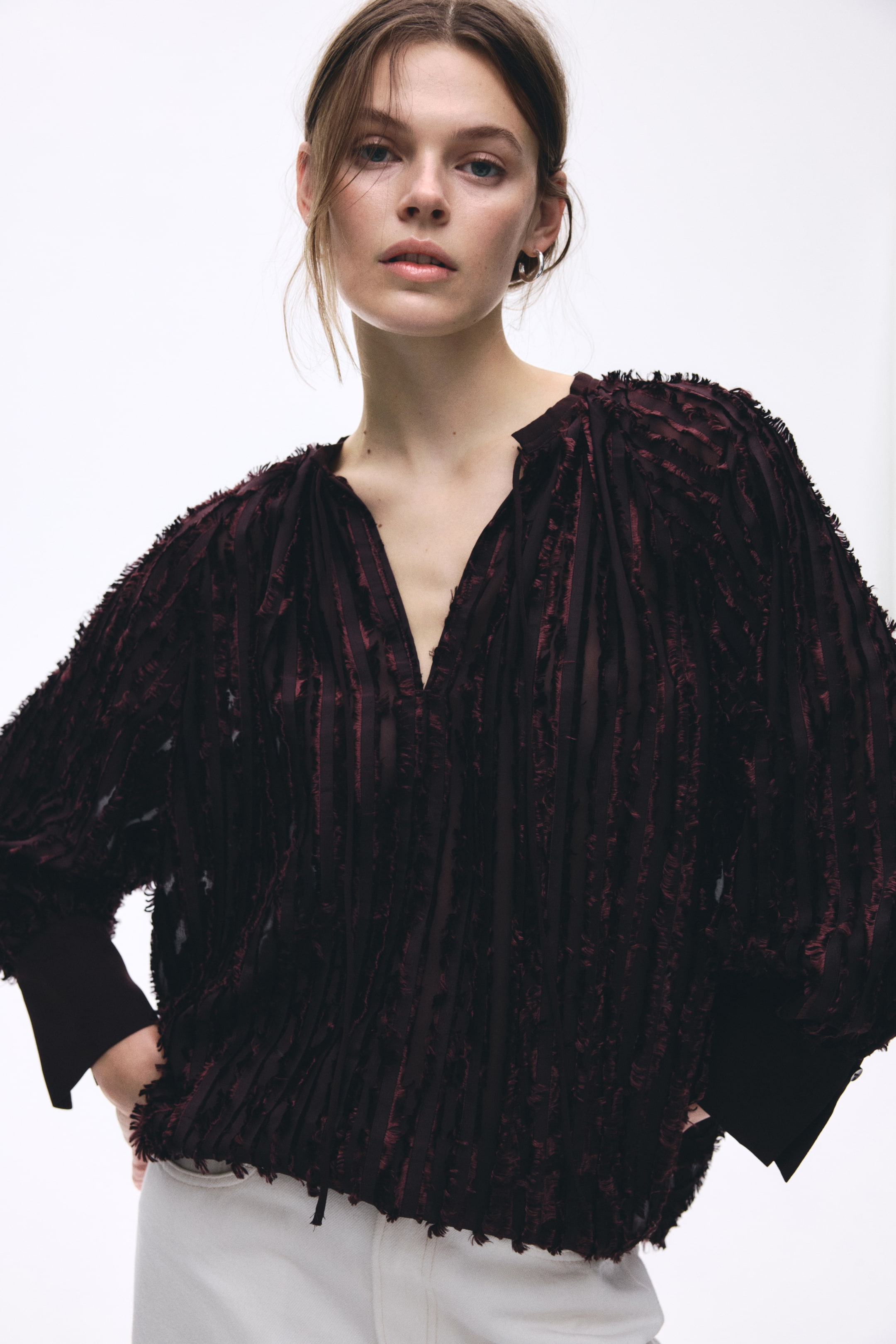 Ver imagen más grande: Un top color burdeos presenta un diseño texturizado con franjas verticales con flecos sobre una tela semi-transparente. Está diseñado con un escote en V dividido y mangas largas que se recogen en puños anchos y ajustados.