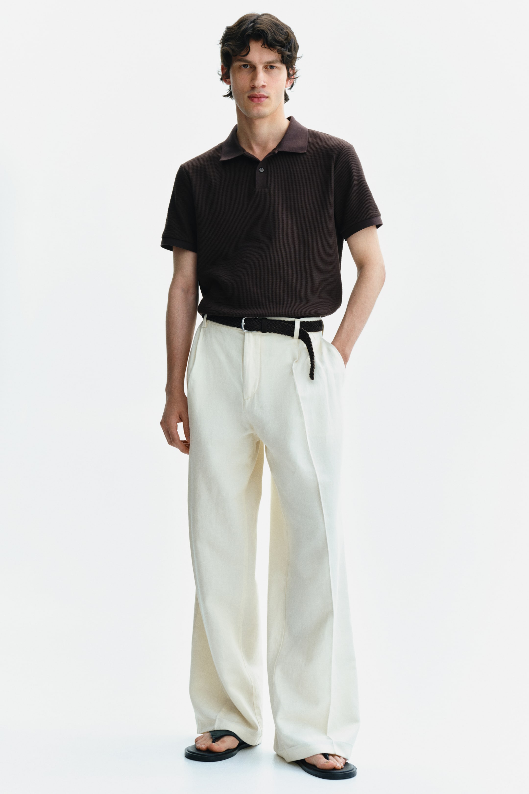 Se større billede: En mørkebrun kortærmet poloshirt med vaffelstrik tekstur, klassisk krave og to-knaps stolpe bæres. Den er stylet nede i lyse beige bukser med brede ben, som har frontlæg og er accessorized med et sort vævet bælte.