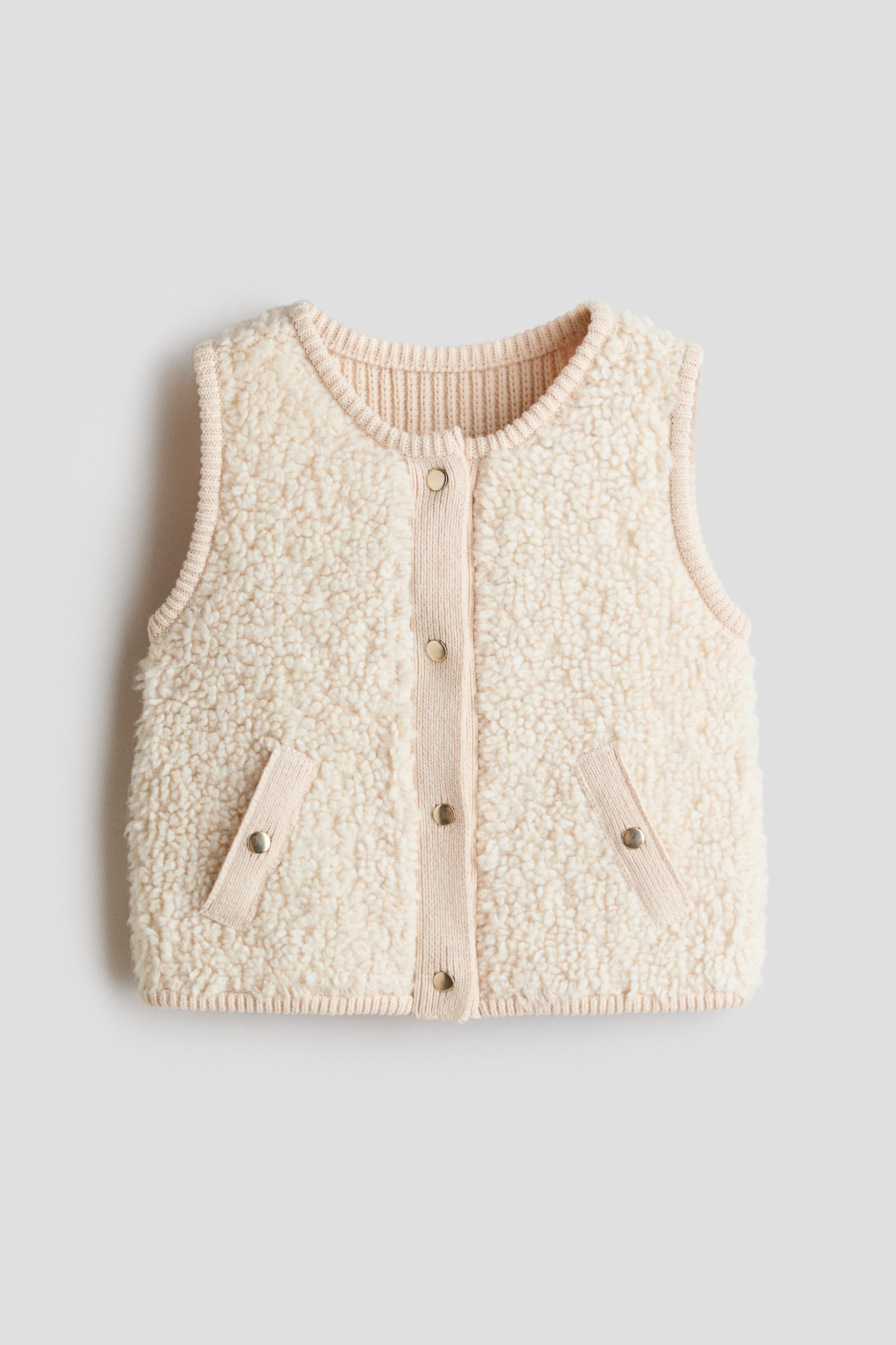 View larger image: A cream-coloured sherpa vest is shown in a front view. It features a ribbed knit round neckline, armholes, and hem, along with a central ribbed knit placket with five snap buttons and two angled patch pockets, each secured with a snap closure.