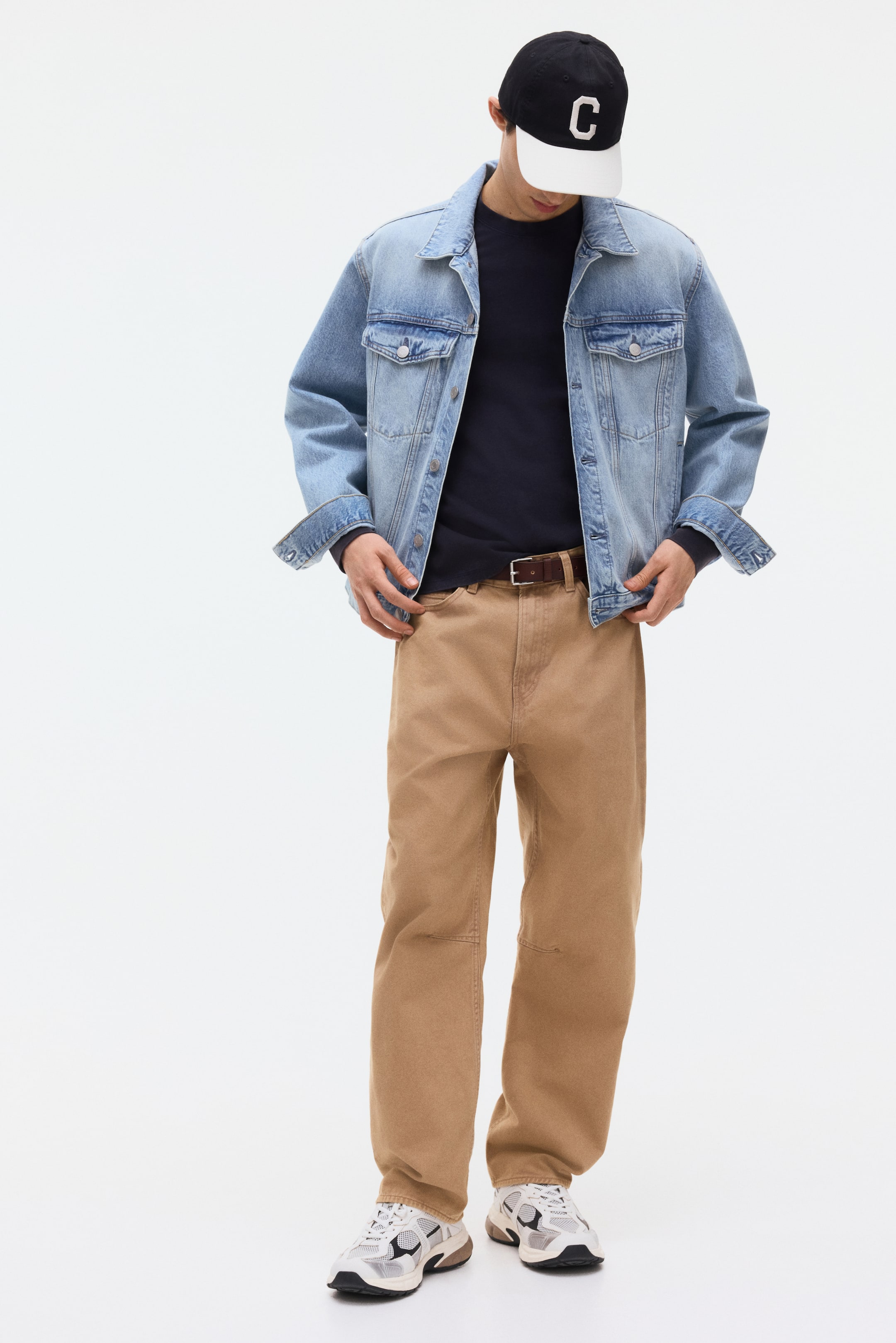 Größeres Bild anzeigen: Eine hellblaue Jeansjacke mit Brusttaschen wird offen über einem dunkelblauen Langarmshirt getragen, kombiniert mit hellbraunen Arbeitshosen im Workwear-Stil mit Knieverstärkungen und einem braunen Ledergürtel. Eine schwarz-weiße Baseballkappe mit einem weißen "C" wird getragen, und weiße, schwarze und beige Sportschuhe sind an den Füßen sichtbar, was eine ganzheitliche, frontale Ansicht des Outfits präsentiert.