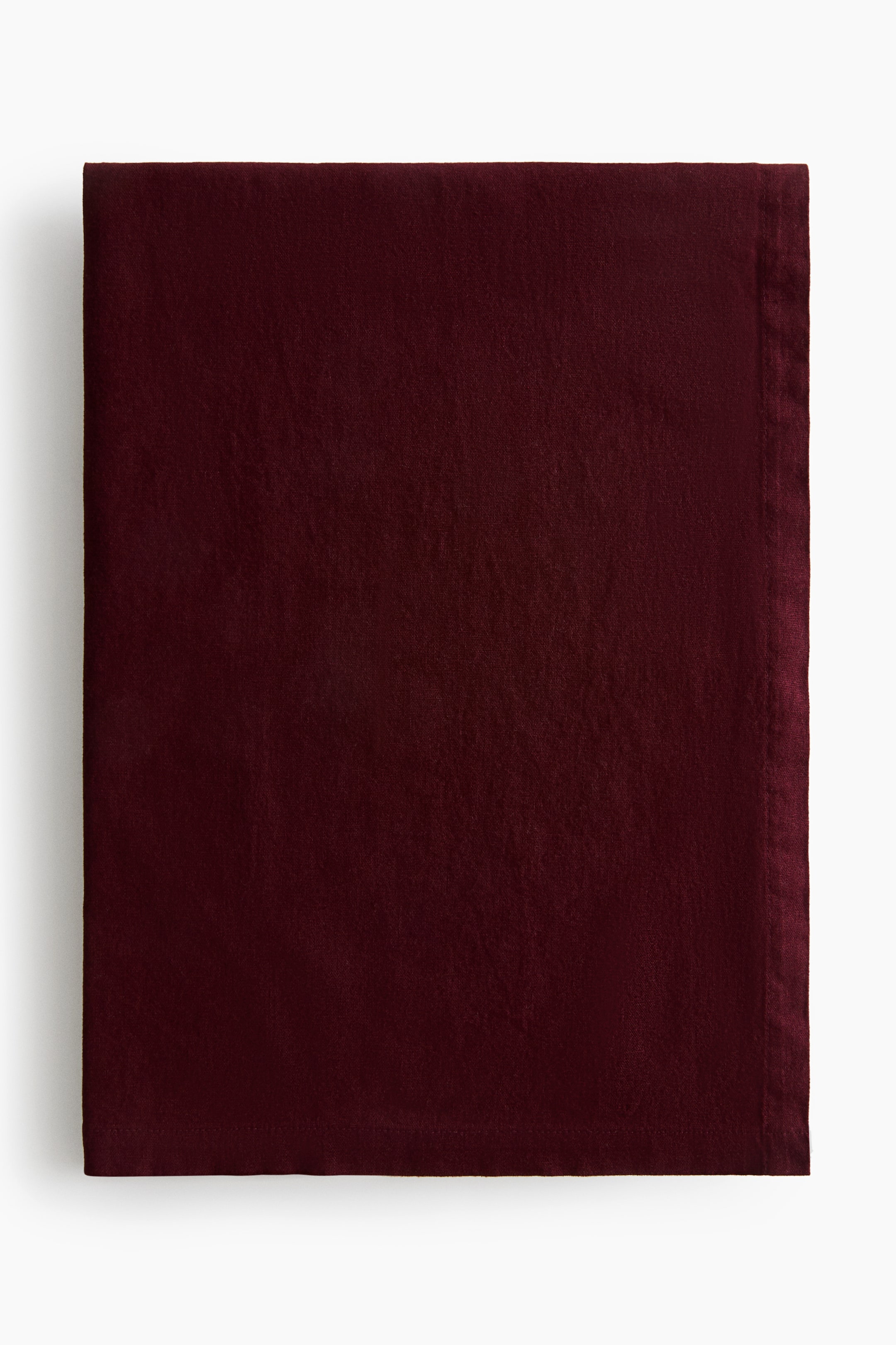 View larger image: A woven tablecloth in a cotton-linen blend is shown folded from the front in a deep burgundy hue. The fabric showcases a textured matte finish and features a visible folded hem along the edges.