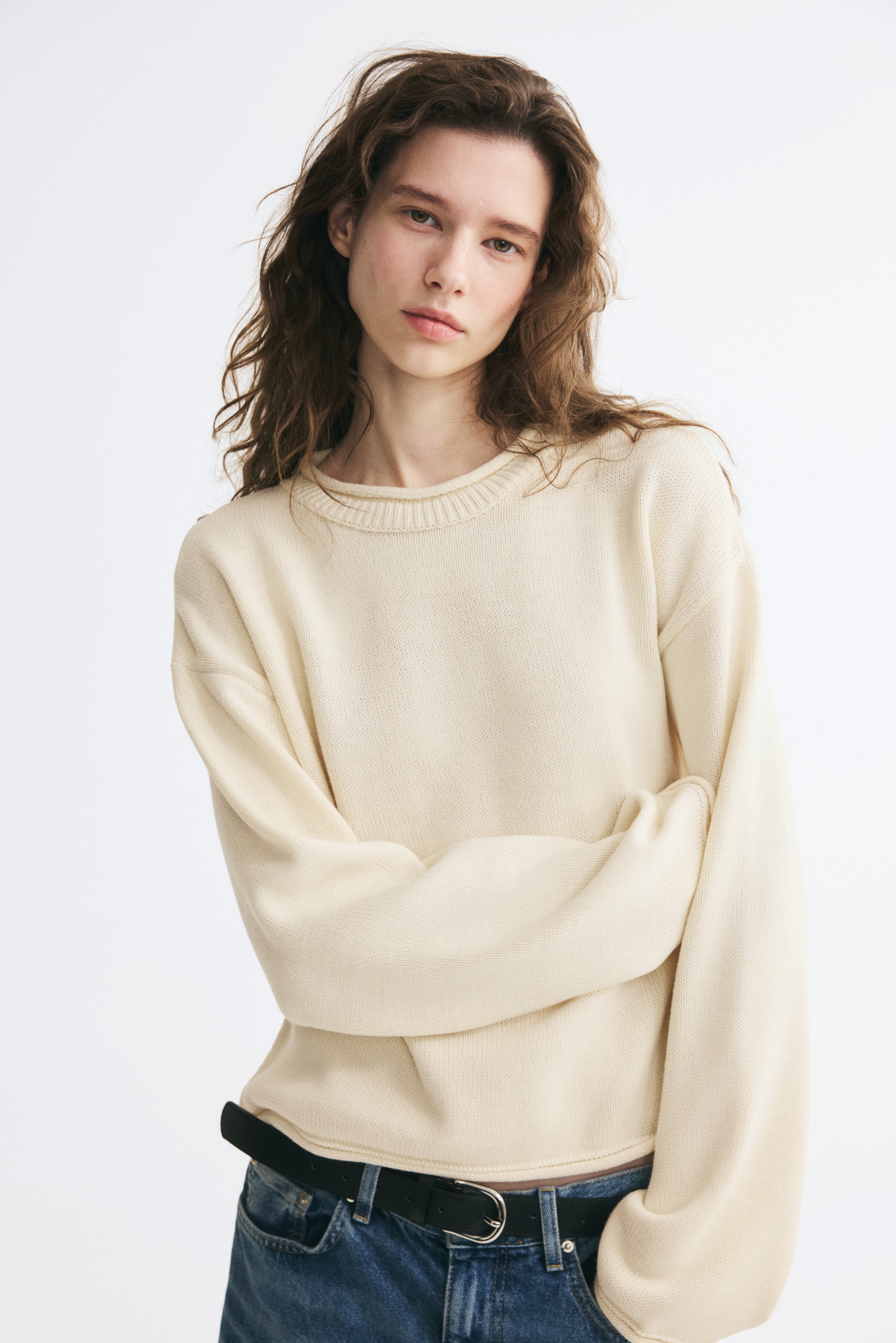 A cream-coloured knitted jumper is hanging facing the front, featuring a ribbed crew neck, dropped shoulders, and long sleeves with rolled cuffs and a matching rolled hem.