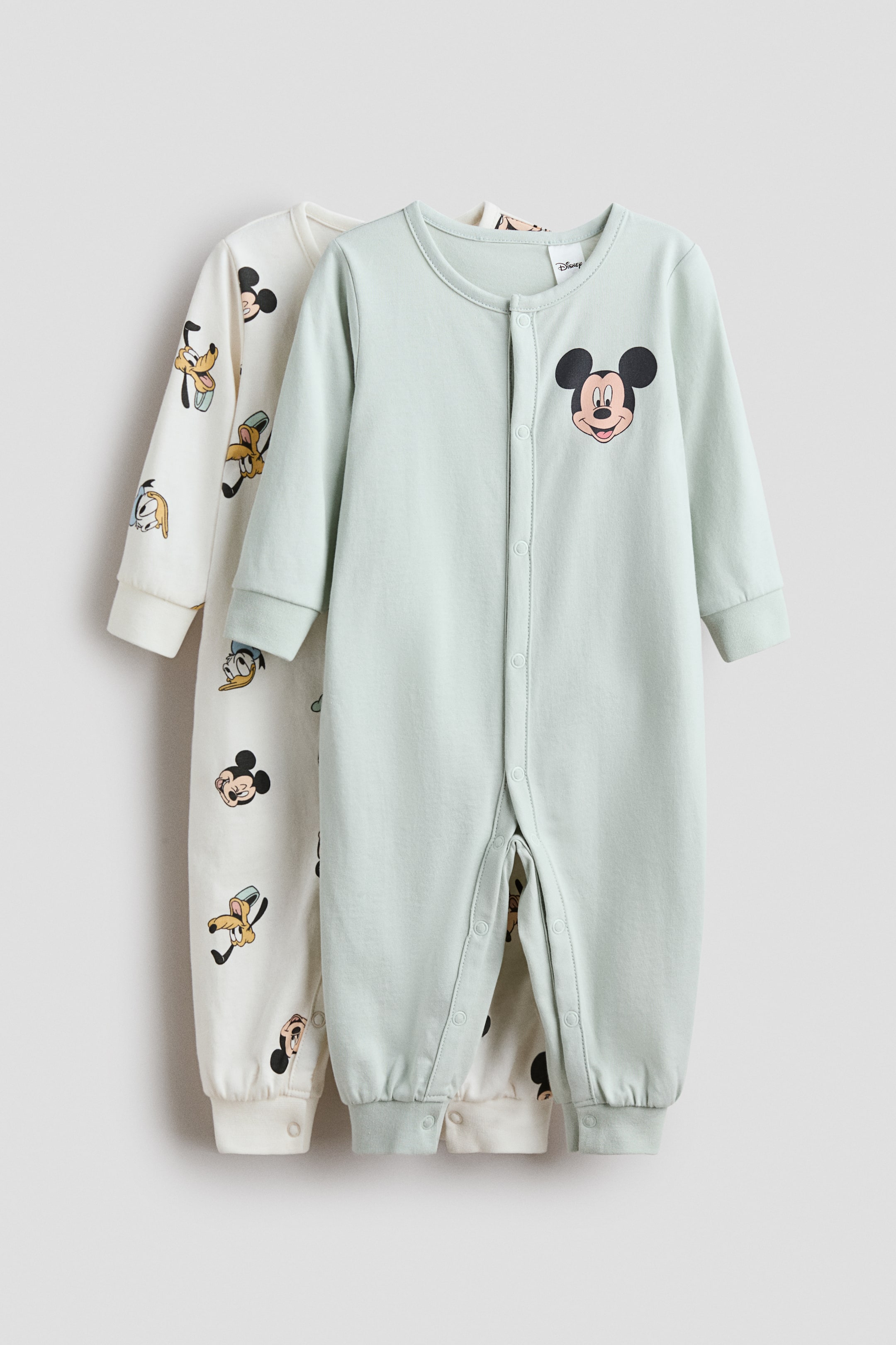 이미지 확대 보기: 두 개의 면 저지 수트가 앞에서부터 제시됩니다. 앞에 있는 수트는 연한 녹색으로, 가슴에 미키 마우스 머리 그래픽이 있고, 앞면과 한쪽 다리에 똑딱이 단추가 있으며, 긴 소매와 골지 커프스 및 밑단이 있습니다. 그 뒤에 부분적으로 보이는 수트는 오프 화이트 색상으로 미키 마우스, 도널드 덕, 구피 머리 전체 프린트가 있으며, 긴 소매와 골지 커프스 및 밑단이 특징입니다.