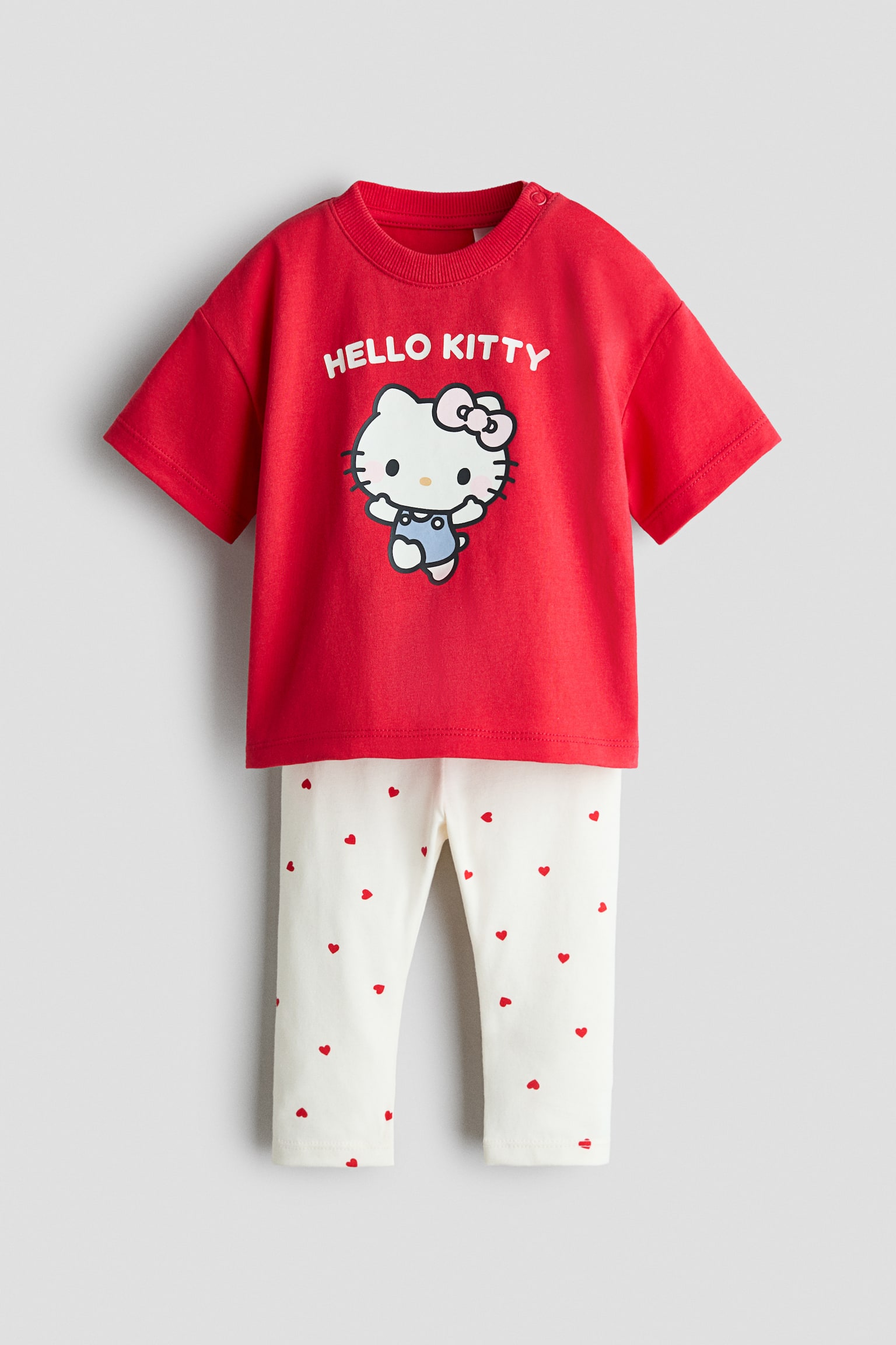 A front view of a two-piece baby outfit is displaying a red short-sleeved t-shirt paired with off-white leggings. The t-shirt is featuring a ribbed crew neck with shoulder snap buttons and a central Hello Kitty graphic under arched white text, while the leggings are showing a scattered pattern of small red hearts.