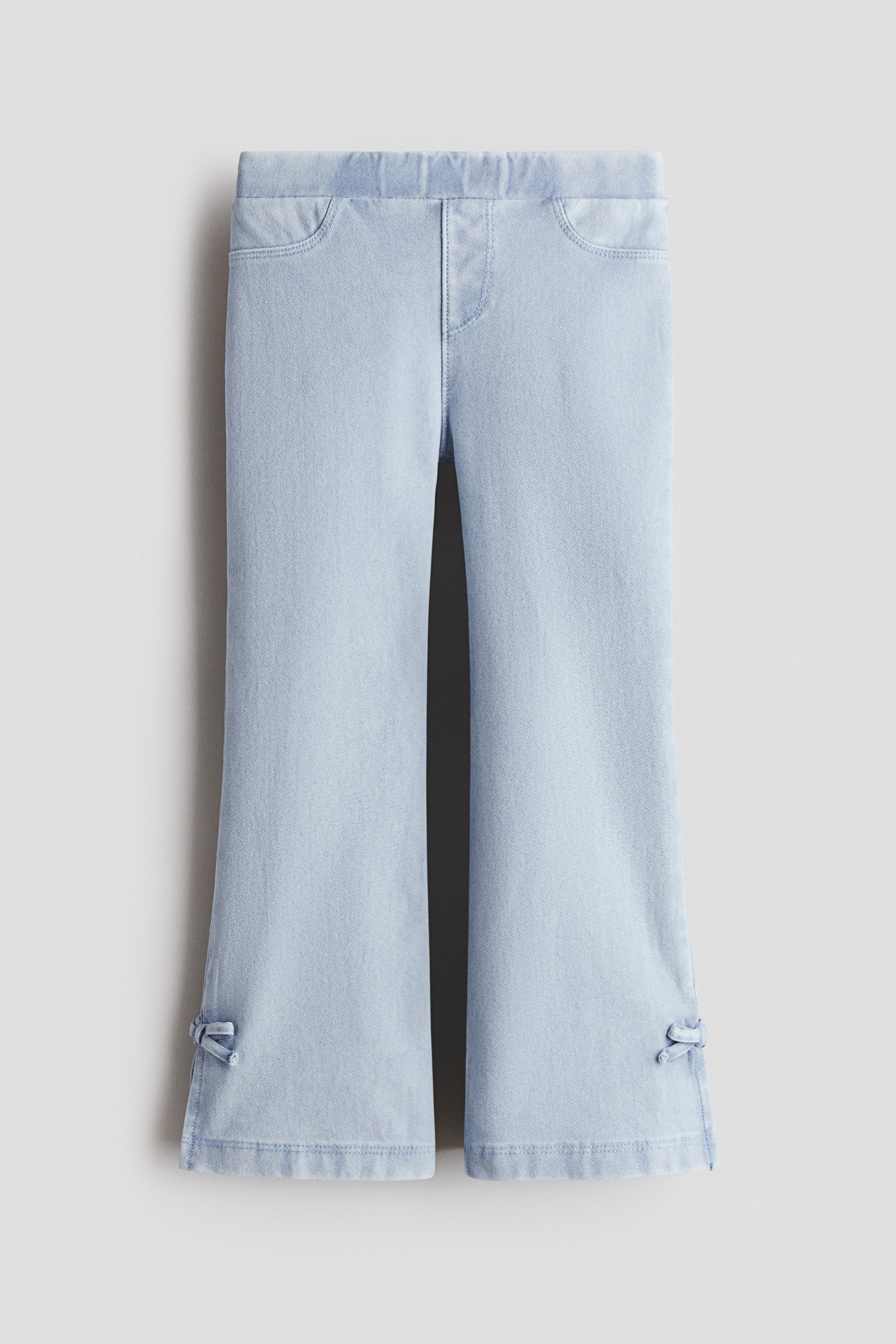 Xem ảnh lớn hơn: Jeggings jersey giả denim màu xanh nhạt được chụp ở mặt trước, có phần eo chun co giãn và phéc-mơ-tuya giả. Các ống loe có túi trước trang trí và được điểm xuyết bằng chi tiết họa tiết giống nơ ở phía ngoài miệng ống mỗi bên.