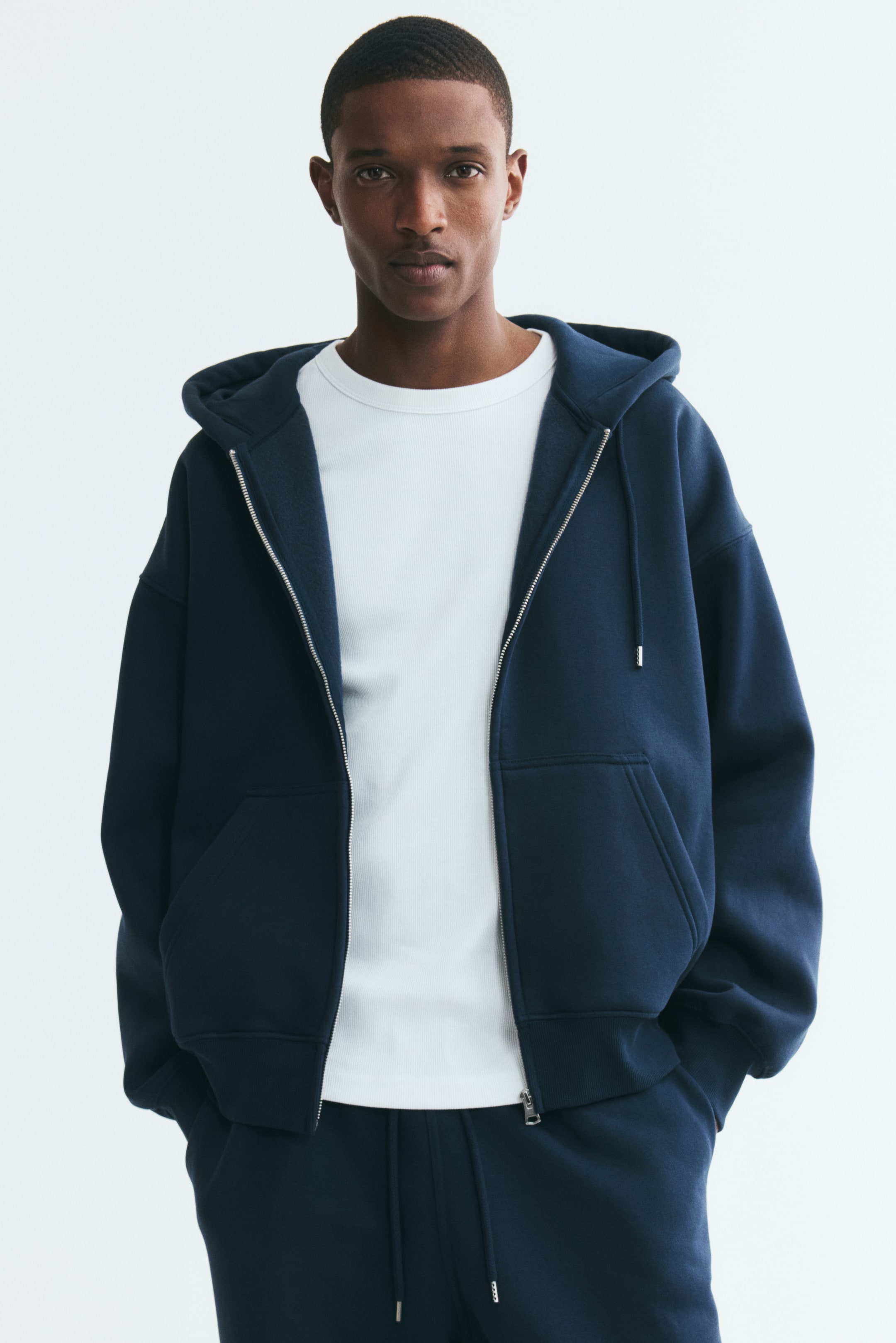 View larger image: A navy blue zip-up hoodie in an oversized fit is worn open by the model over a white crewneck T-shirt. The hoodie, which features dropped shoulders, front pockets, and a drawstring hood, is paired with matching navy sweatpants.