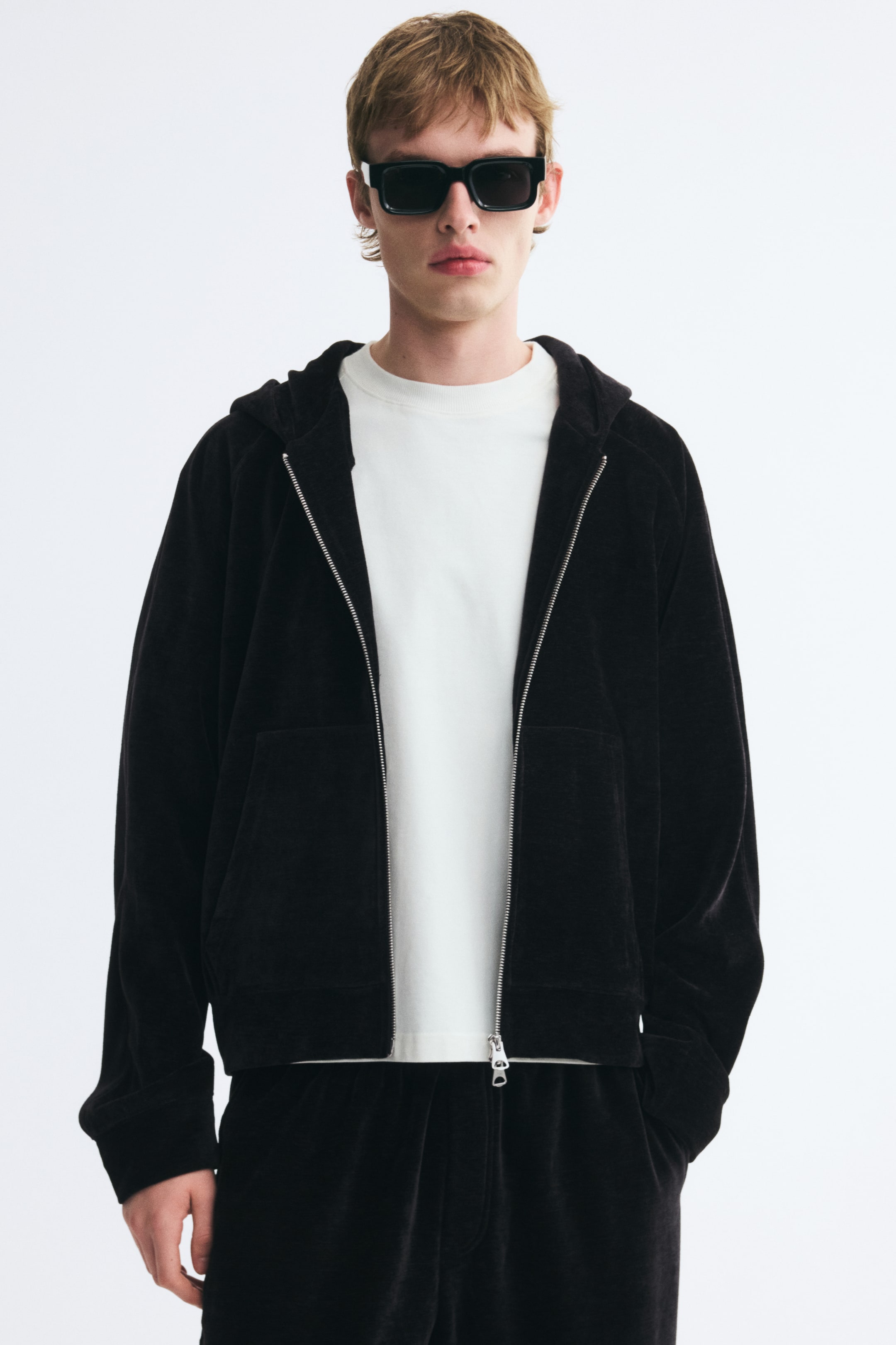 View larger image: A black velour hooded jacket is worn open, featuring a silver front zipper, front pockets, and a relaxed fit with dropped shoulders. The jacket is layered over a white crew-neck T-shirt and paired with matching black velour trousers on the model.
