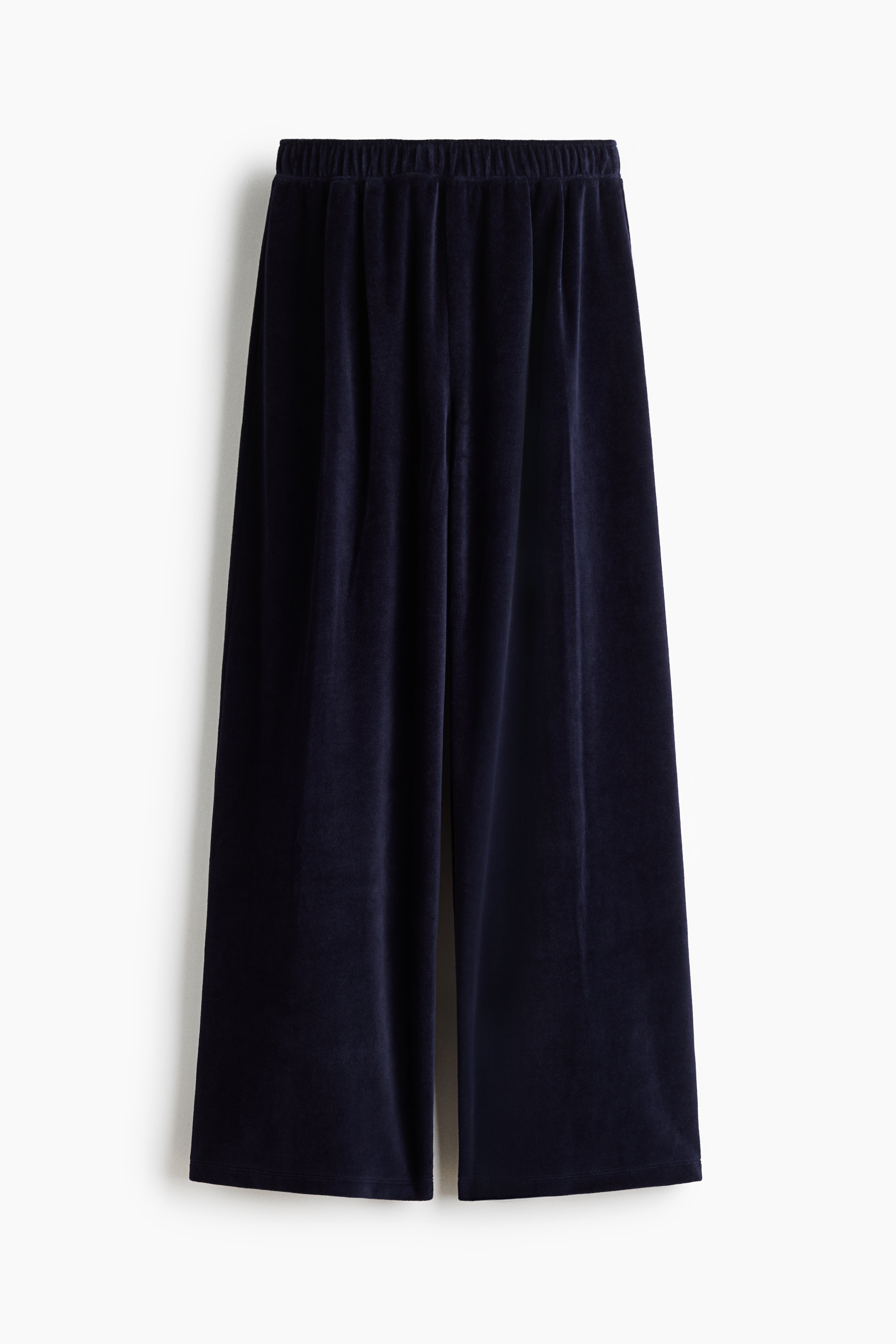 The model is wearing dark blue velour joggers with an elasticated waistband and straight legs featuring pleats at the top. The joggers are styled with a matching open velour zip-up jacket and a white tank top.