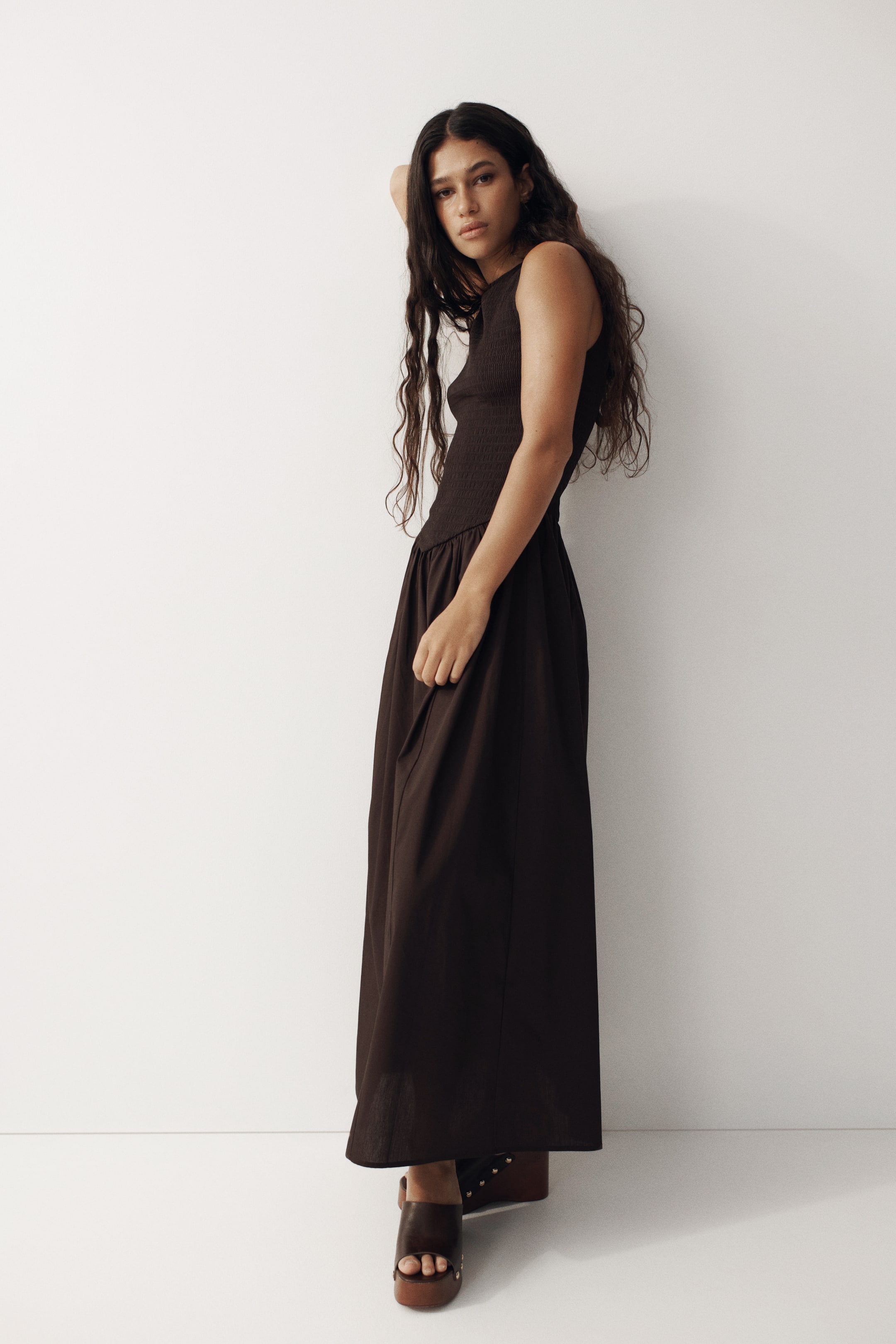 View larger image: A dark brown maxi dress features a fitted, smocked sleeveless bodice with a round neckline, flowing into a smooth, full-length skirt, angled to the left to showcase its silhouette.
