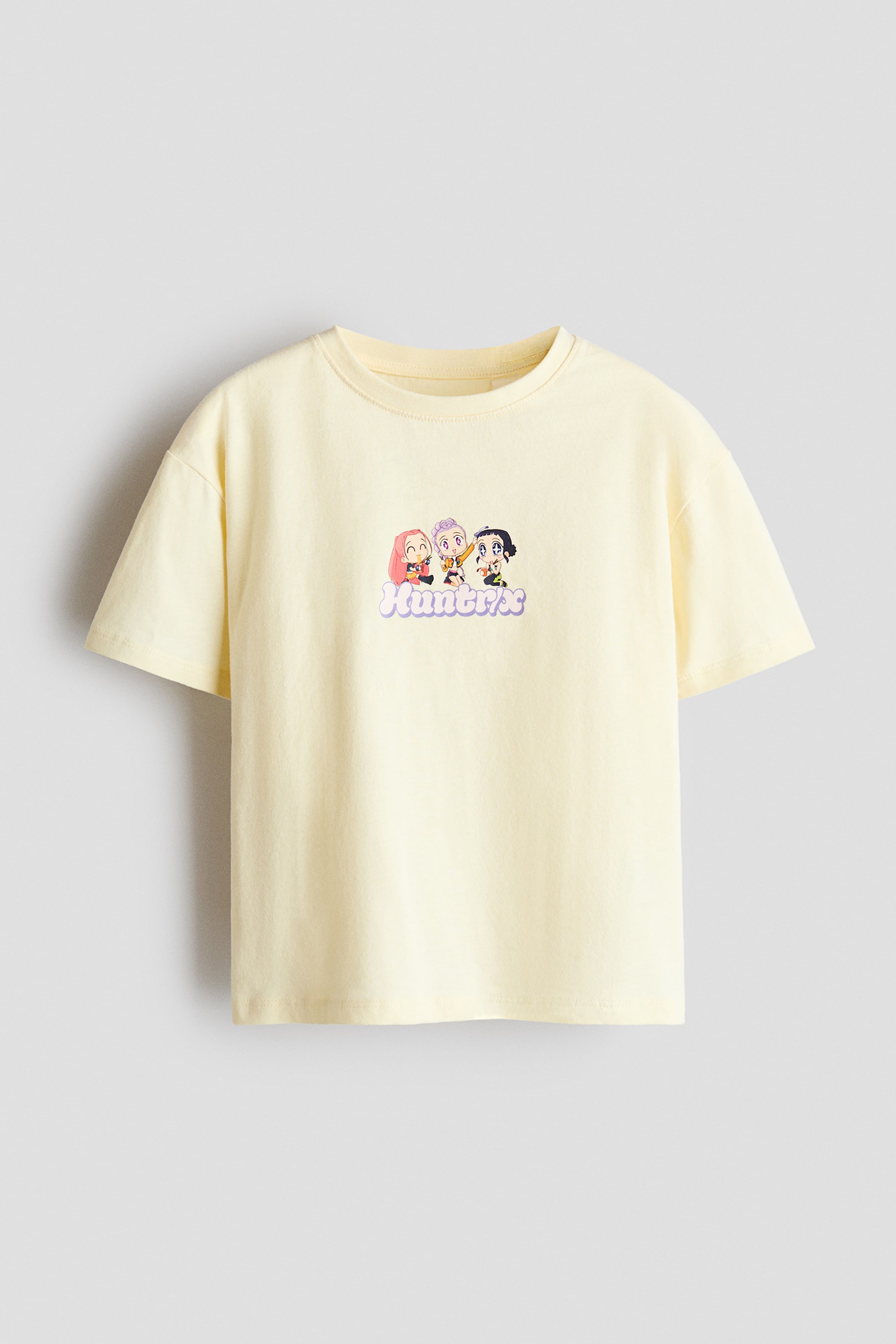 View larger image: A light yellow cotton jersey T-shirt is shown from the front, featuring a rib-trimmed crew neckline, dropped shoulders, and short sleeves. The center chest features a small graphic print depicting three cartoon characters positioned above the stylized purple word "Huntrix."