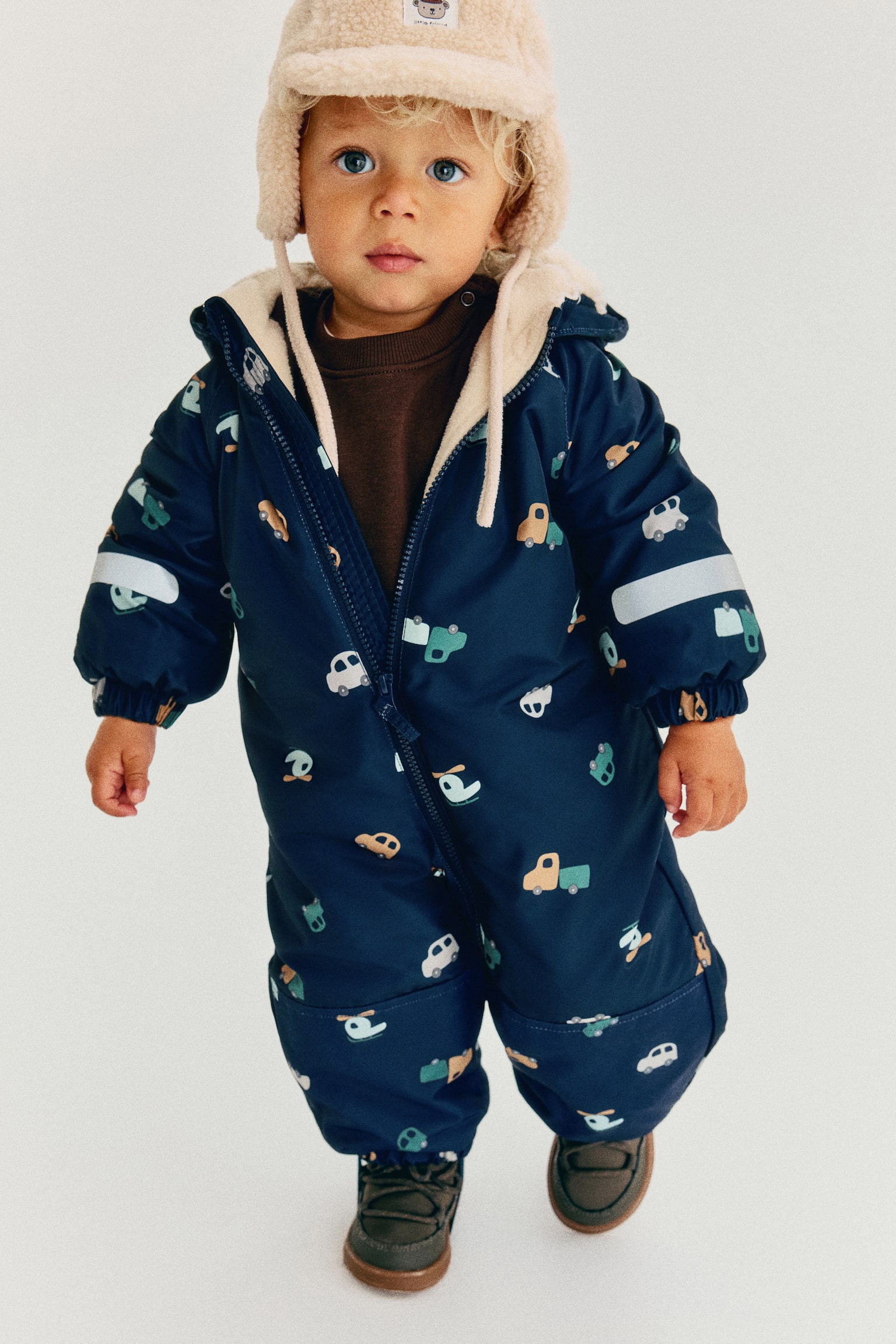 Ein marineblauer Schneanzug für Kleinkinder mit einem Allover-Print aus kleinen Autos und Hubschraubern ist von vorne zu sehen und zeigt durch den teilweise geöffneten ReiÃverschluss vorne ein cremefarbenes Fleecefutter. Der Schneanzug ist mit einer angesetzten Kapuze, elastischen Bündchen und waagerechten Reflektorstreifen an den Ãrmeln ausgestattet. - 1