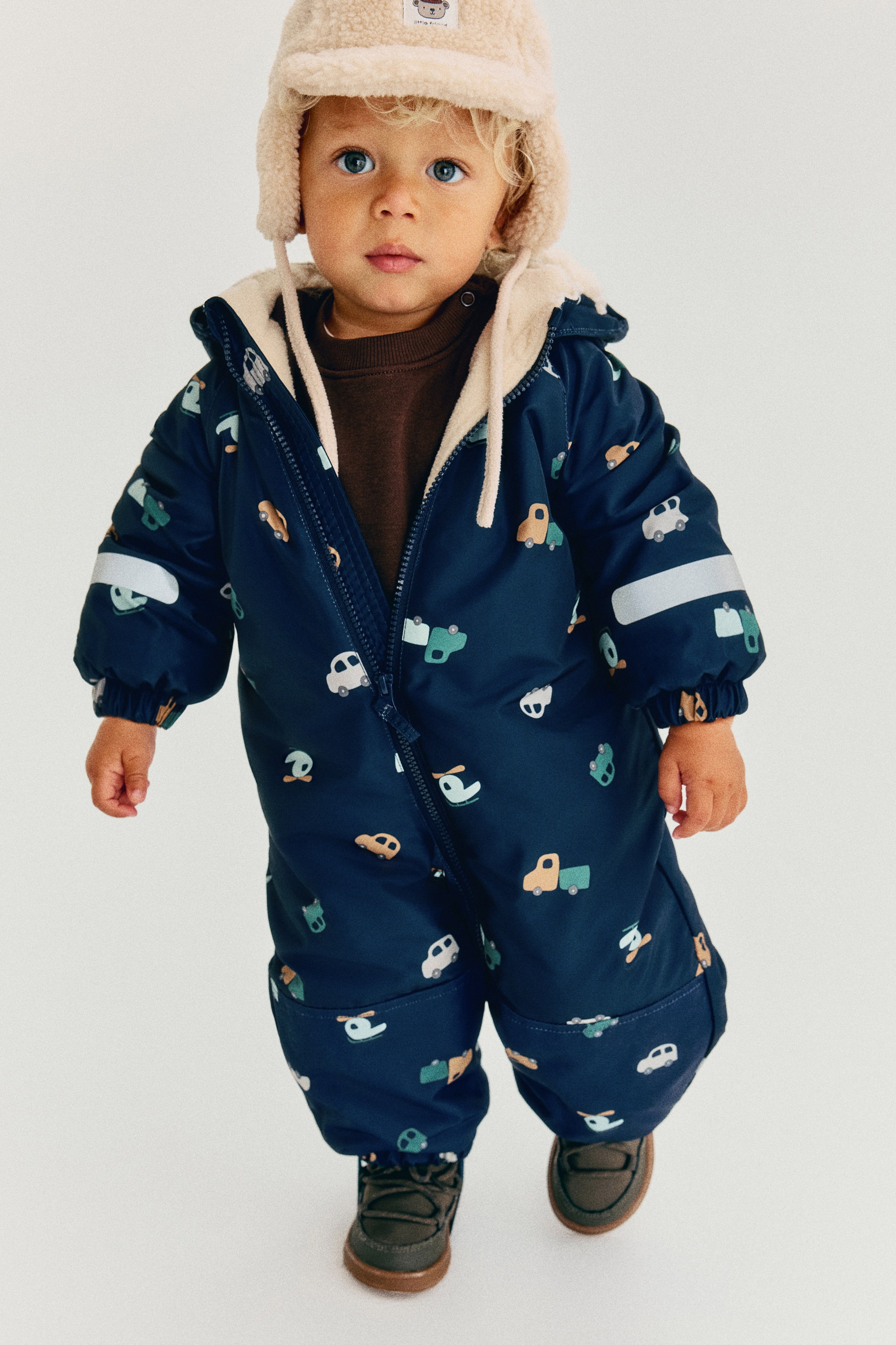 View larger image: A navy blue toddler snowsuit featuring an all-over print of small cars and helicopters is shown from the front, revealing a cream-coloured fleece lining through its partially unzipped front closure. The snowsuit is designed with an attached hood, elasticized cuffs, and horizontal reflective strips on the sleeves.