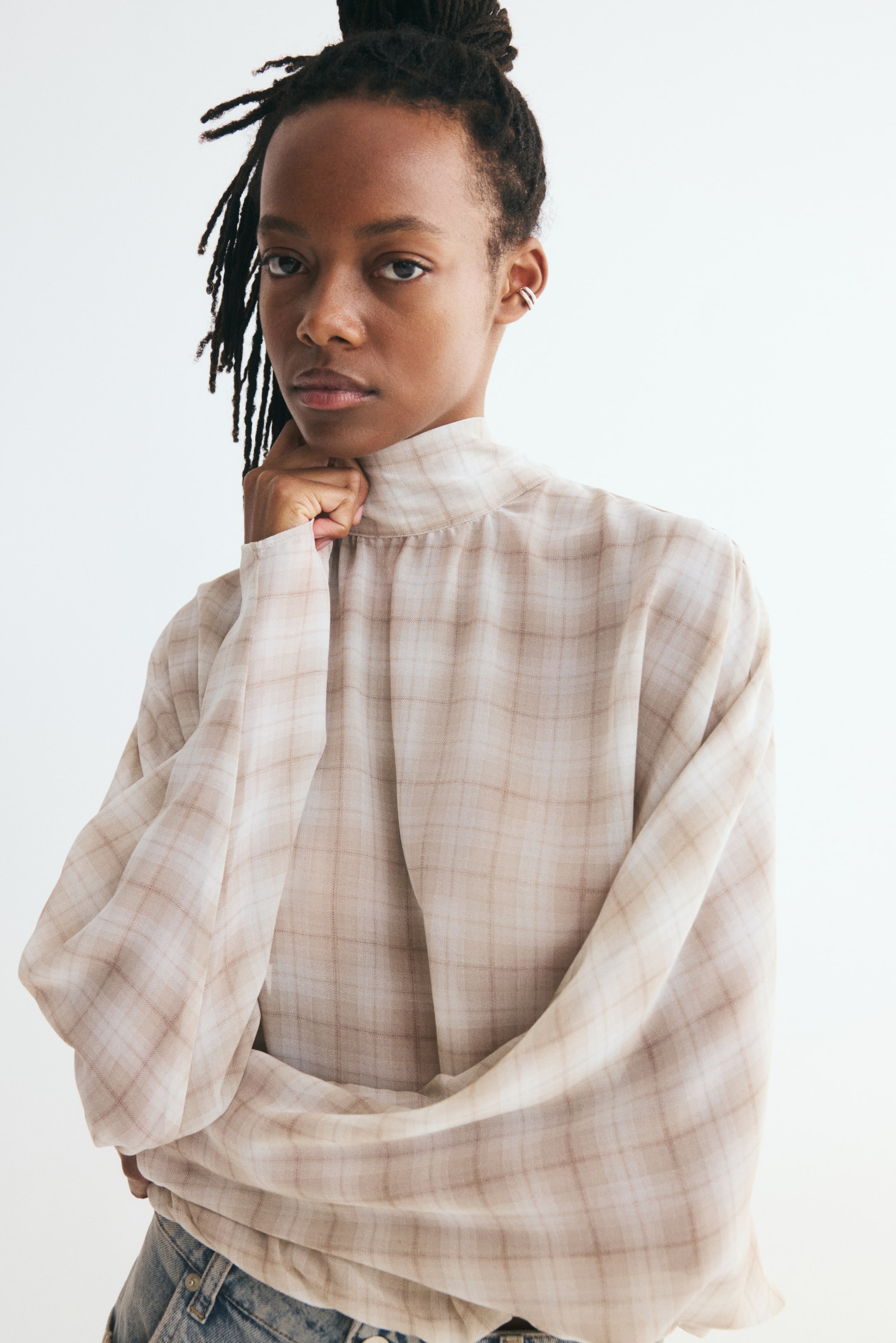 View larger image: A tie-back stand-collar blouse in a beige plaid pattern is draping loosely in a front-facing view. The blouse features a high, gathered stand collar and long, relaxed sleeves tucked into denim jeans.