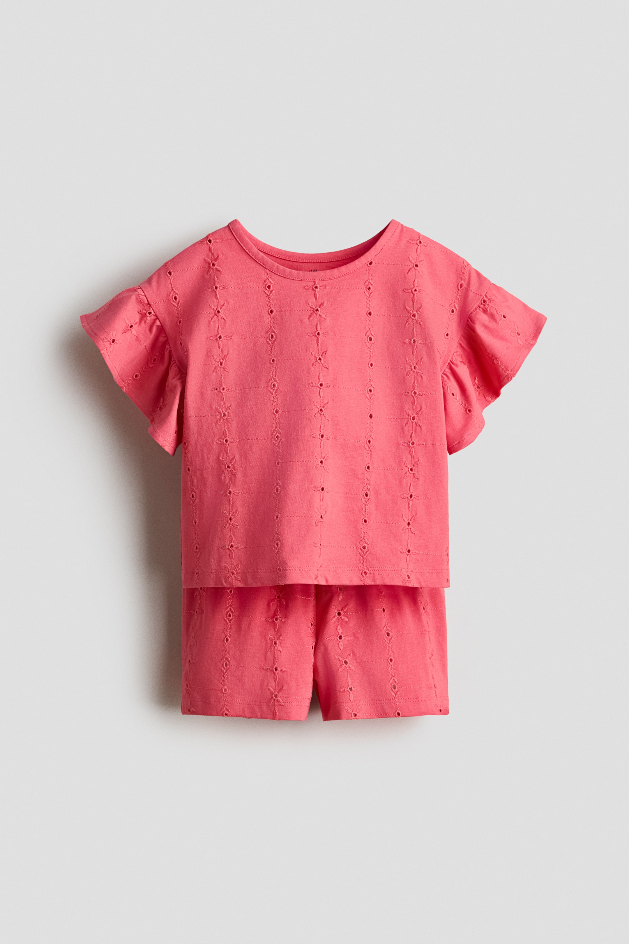 View larger image: A coral cotton jersey set includes a top and matching shorts, both adorned with broderie anglaise. The top is displayed from the front, featuring a narrowly trimmed round neckline, dropped shoulders, and flutter sleeves. The shorts are styled with an elasticated paper bag waist.