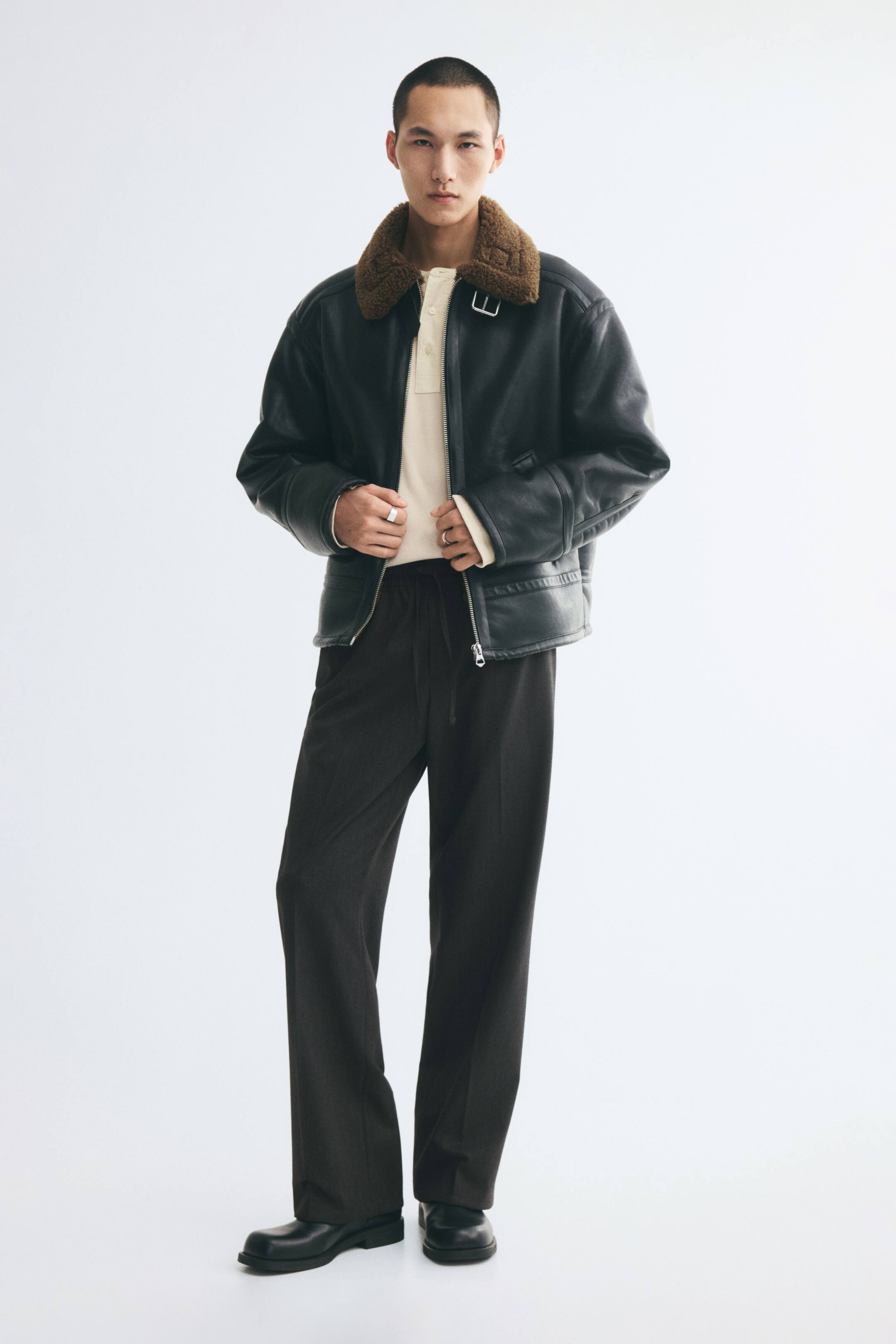 View larger image: Tailored joggers in a dark, woven fabric are worn by the model, featuring a relaxed fit, an elasticated drawstring waist, and visible creases down the front of the legs. The joggers are styled with a cream-coloured polo shirt, a black leather jacket with a brown shearling collar, and black leather shoes.