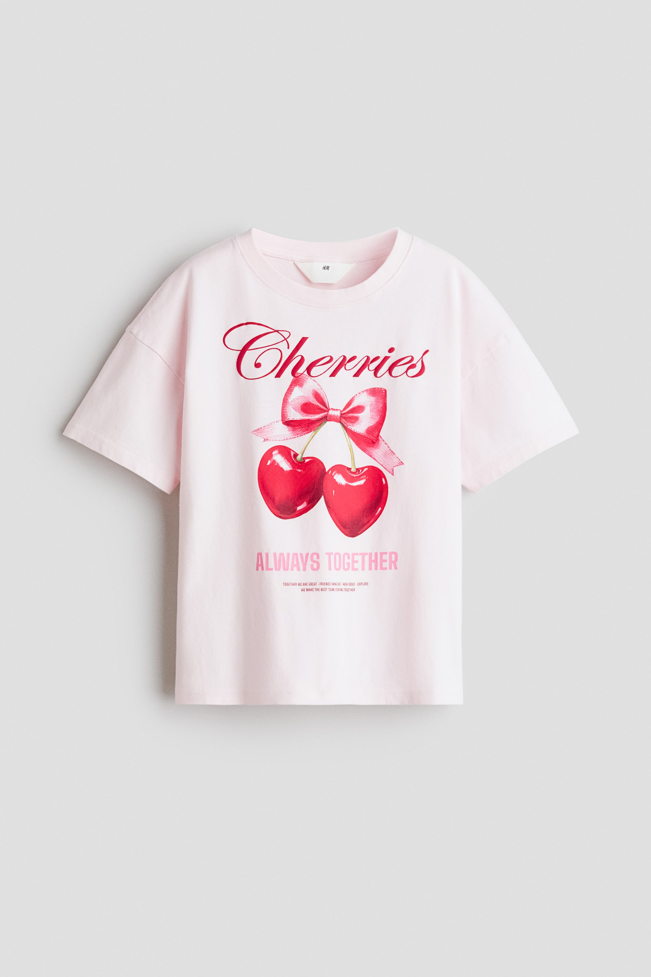 Größeres Bild anzeigen: Ein hellrosa bedrucktes Baumwolljersey-T-Shirt mit rundem, geripptem Ausschnitt und überschnittenen Schultern. Auf der Vorderseite ist eine Grafik von „Cherries“ in roter Schrift über einer roten Schleife und zwei herzförmigen roten Kirschen zu sehen, darunter sind „ALWAYS TOGETHER“ in pinken Blockbuchstaben gedruckt.
