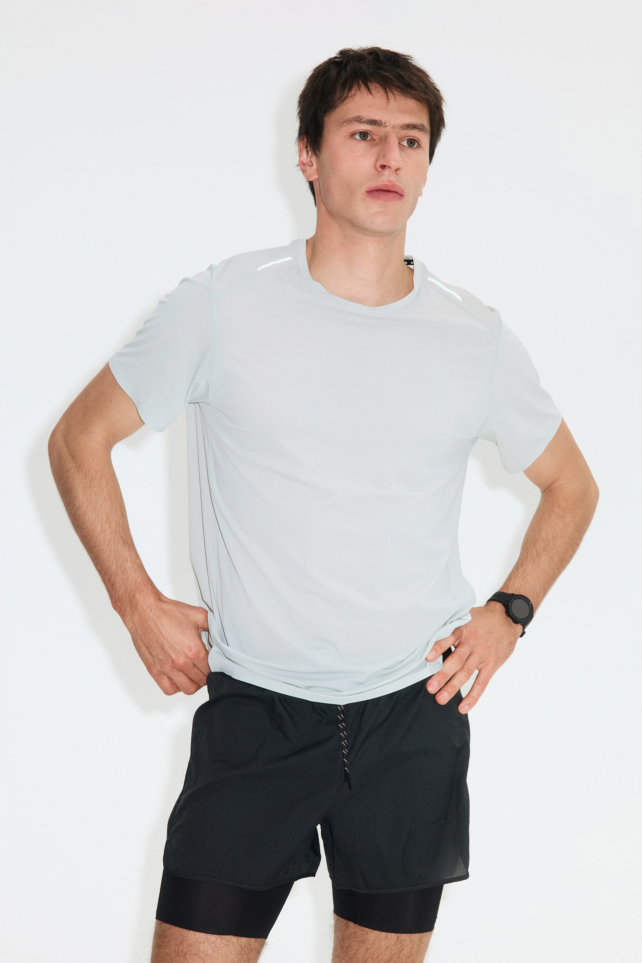 View larger image: The light grey short-sleeved t-shirt showcases a subtle vertical stripe texture and white reflective accents on the shoulders. The black 2-in-1 shorts feature a crinkled outer layer with a speckled drawstring and a longer, ribbed inner compression layer.