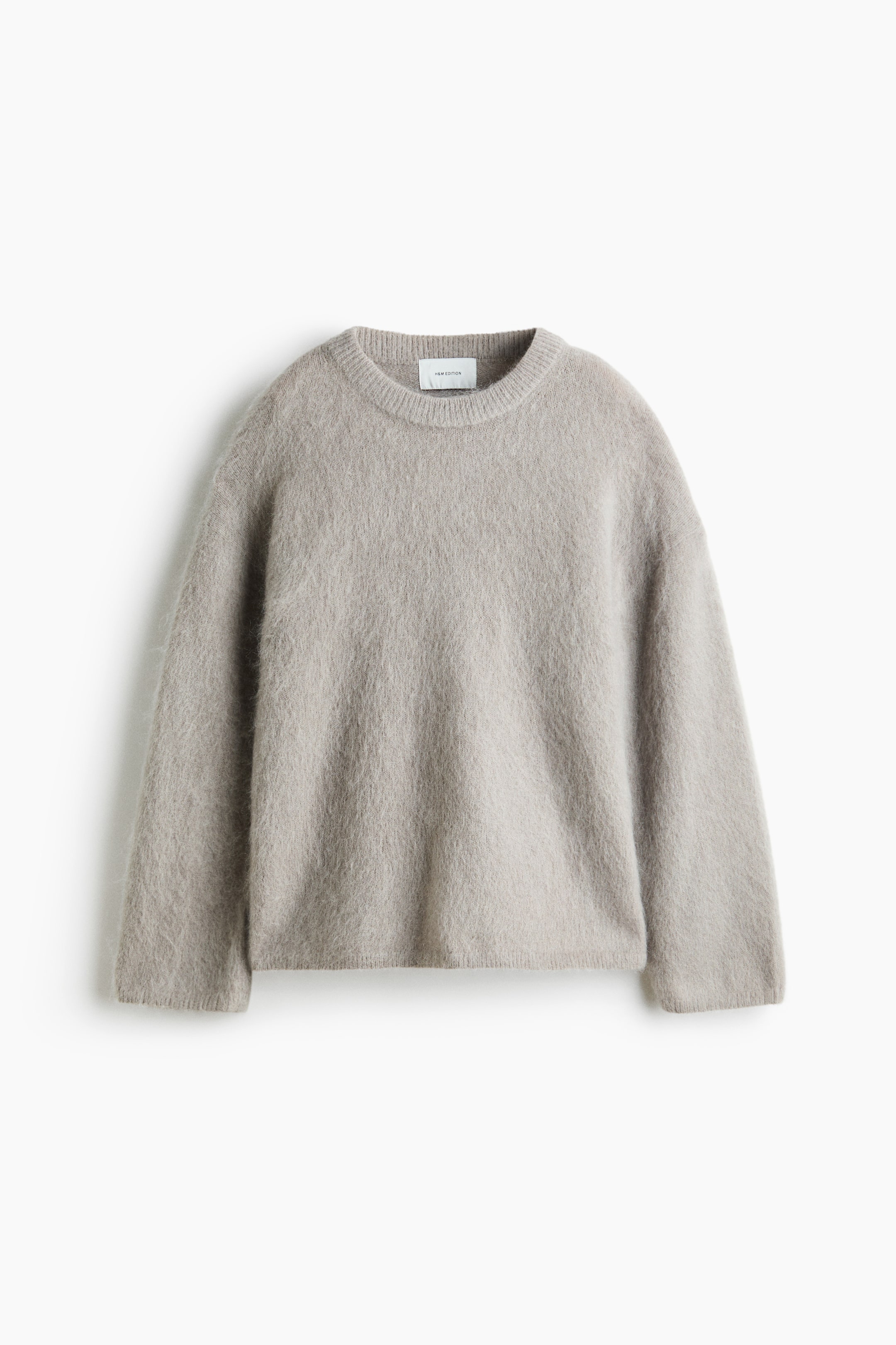 Visa större bild: En oversized jumper visas framifrån och har en mjuk, ljusgrå stickad struktur, rund halsringning, nedsänkta axlar, långa ärmar samt ribbning vid halsringningen, ärmsluten och nederkanten.