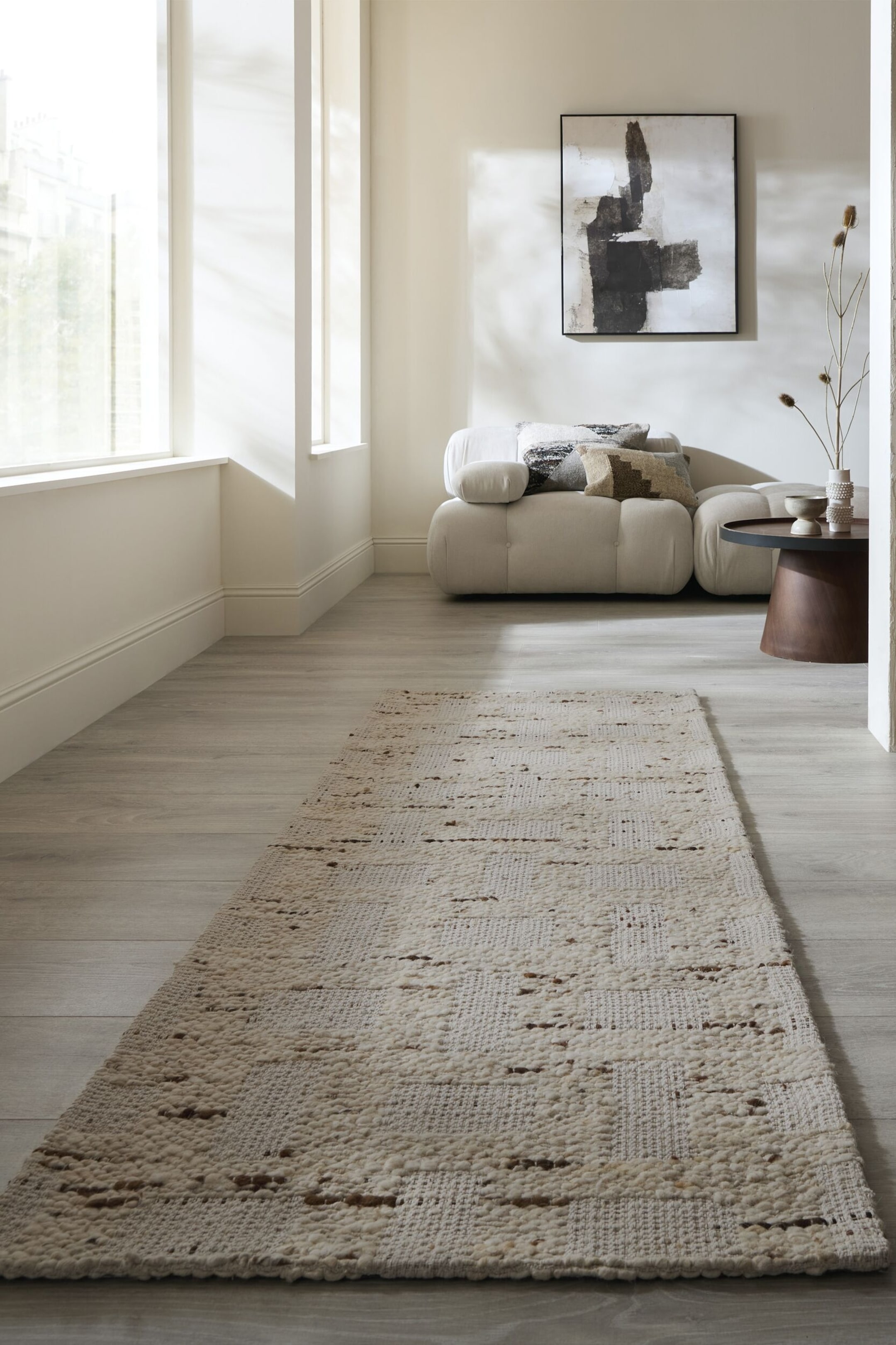 Visa större bild: En lång, rektangulär löpare ligger vinklad på ett ljust golv, med ett off-white och krämfärgat texturerat vävt mönster. Den visar oregelbundna horisontella linjer av mörkare fibrer och alternerande plattare väv med upphöjda, tuftade sektioner.