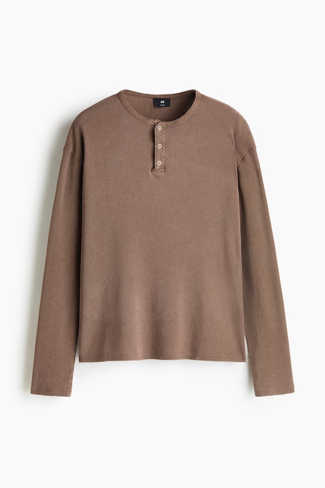 Een bruin Henley-top met lange mouwen van wafelstructuur katoenjersey is van voren afgebeeld, met een relaxed fit en verlaagde schouders. De top heeft een ronde halslijn met een knoopsluiting met drie knopen, eenvoudige rechte manchetten en een recht afgesneden zoom. - 1