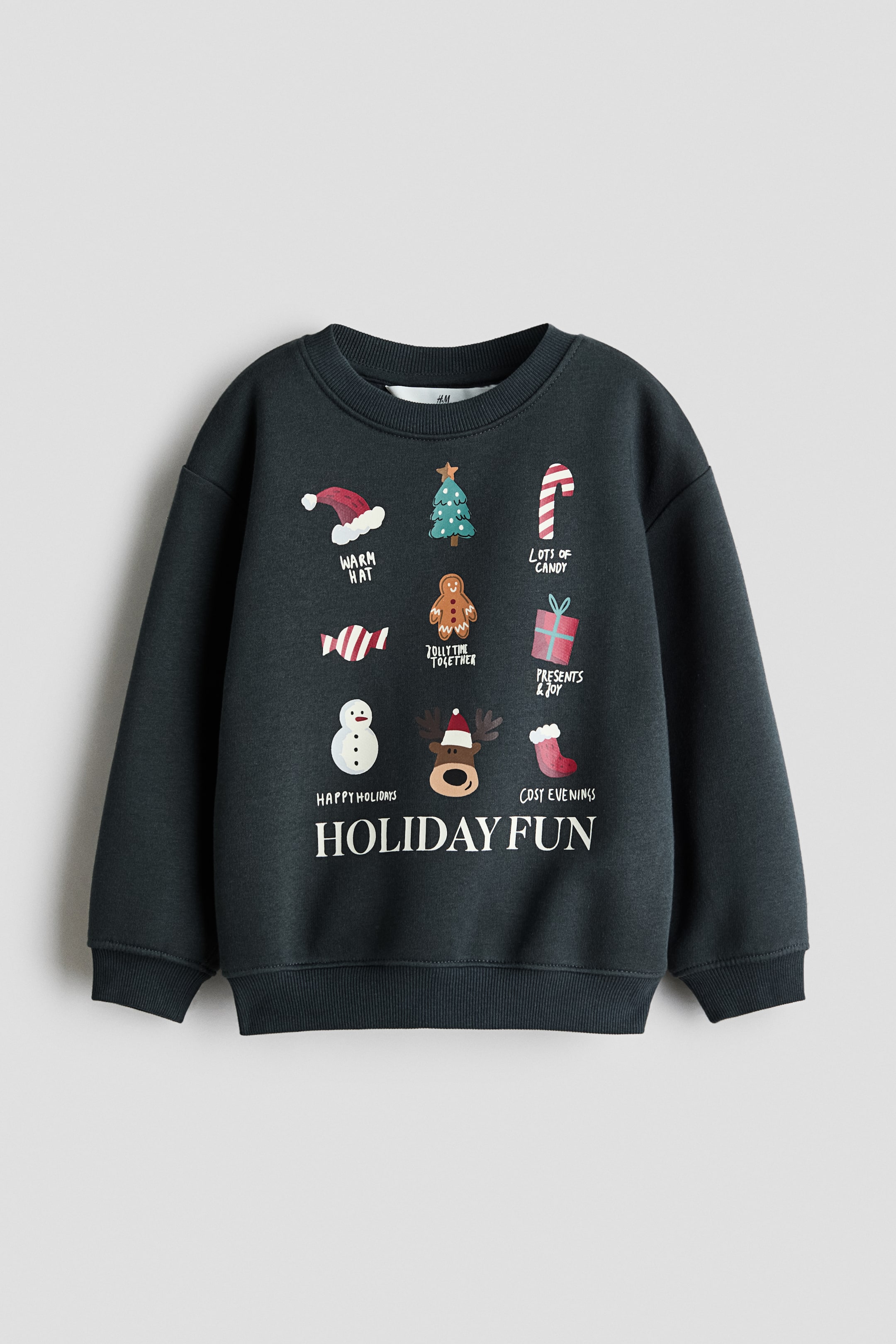 Visa större bild: Denna mörkgrå sweatshirt visas framifrån och har en rund ribbad halsringning, nedhasade axlar, långa ärmar med ribbade muddar och en ribbad fåll. Framsidan visar ett tryckt rutnät av festliga illustrationer och text med högtidstema, med orden ”HOLIDAY FUN” tryckta i vita versaler längst ner.
