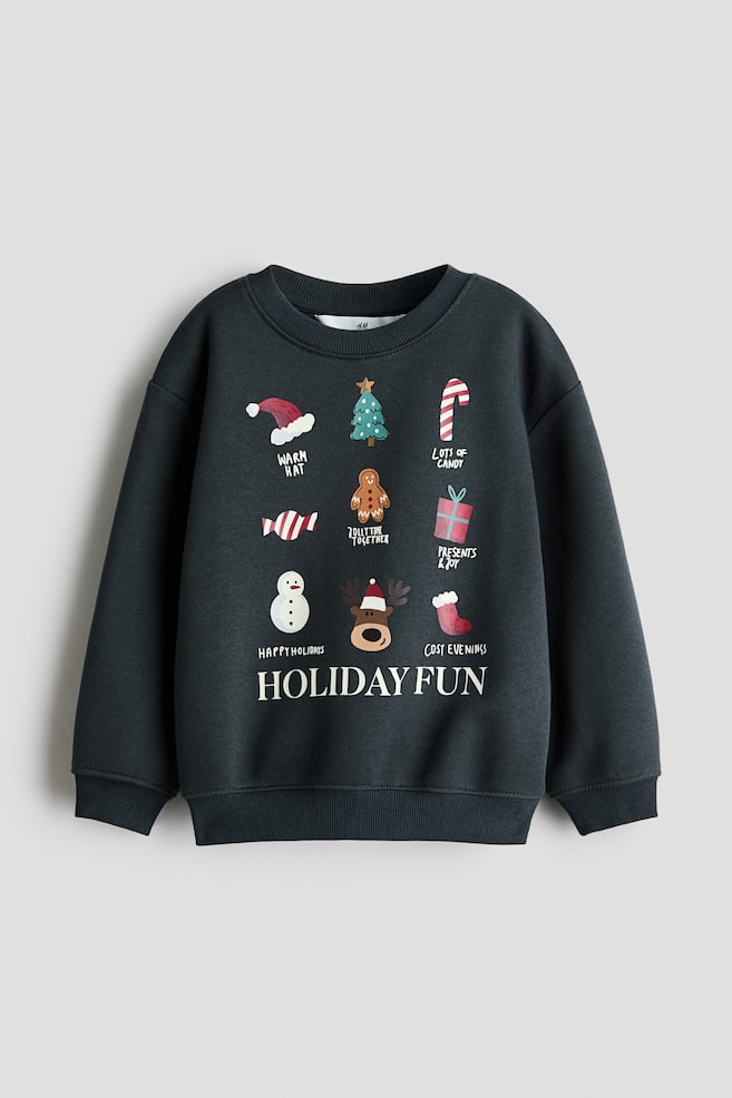 Denna mÃ¶rkgrÃ¥ sweatshirt visas framifrÃ¥n och har en rund ribbad halsringning, nedhasade axlar, lÃ¥nga Ã¤rmar med ribbade muddar och en ribbad fÃ¥ll. Framsidan visar ett tryckt rutnÃ¤t av festliga illustrationer och text med hÃ¶gtidstema, med orden âHOLIDAY FUNâ tryckta i vita versaler lÃ¤ngst ner. - 1