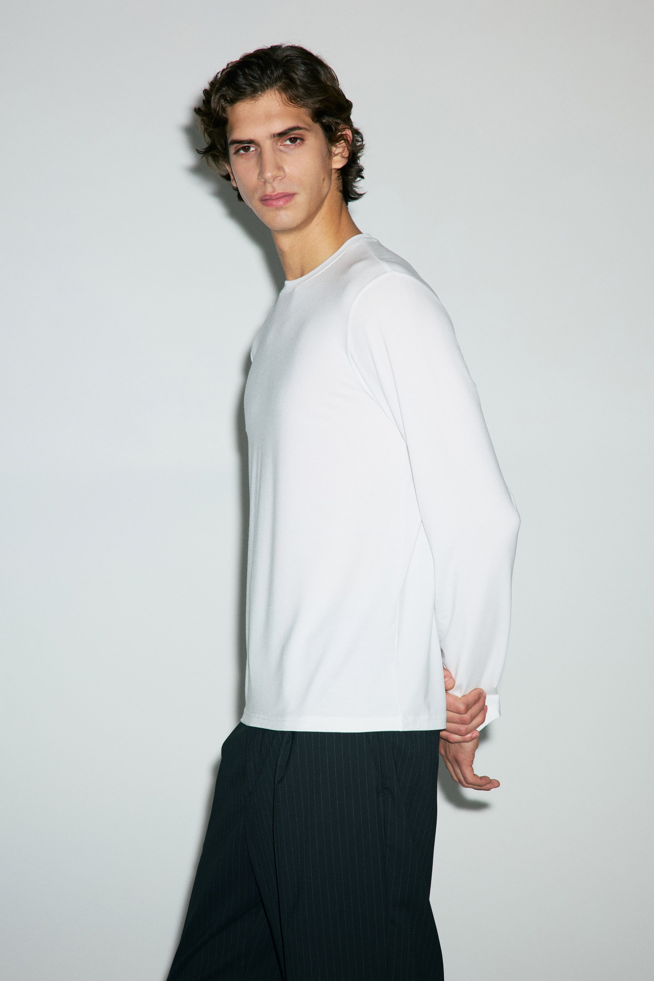 View larger image: A white long-sleeved crew-neck top features a smooth texture and a straight hem, offering a relaxed fit. Black trousers showcase subtle vertical pinstripes and side pockets.