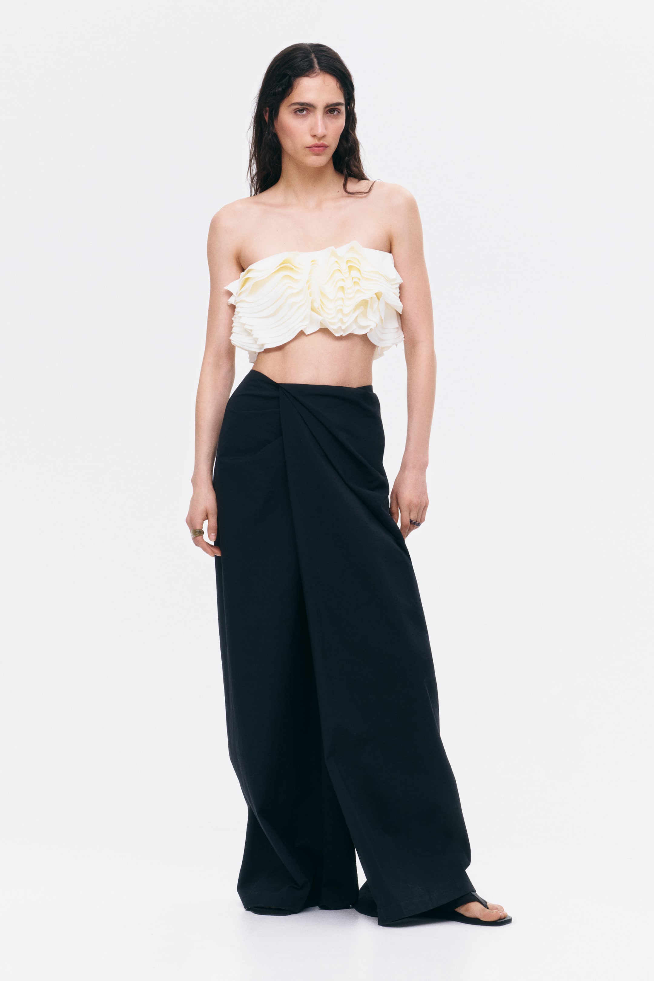 View larger image: This cream strapless crop top showcases layered ruffles elegantly draped across the front. Complementing it are black wide-leg trousers, distinguished by a unique twisted drape detail at the front waist.