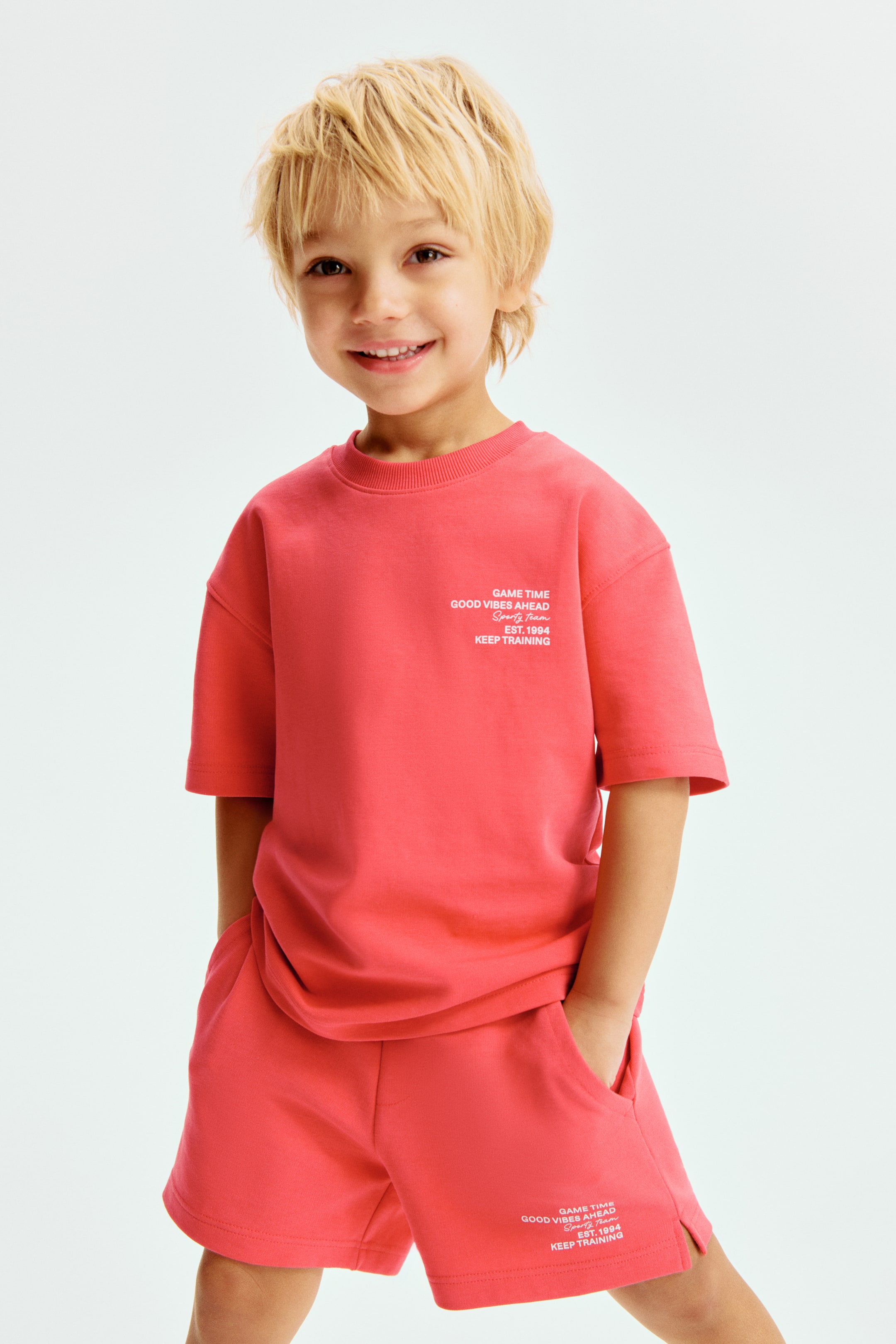 Grösseres Bild ansehen: Ein korallenfarbenes T-Shirt mit kurzen Ärmeln und passende Shorts bilden ein Set, das aus einem leichten Frontwinkel betrachtet wird. Das T-Shirt zeigt einen gerippten Rundhalsausschnitt und weißen Text auf der linken Brust, während die Shorts mit Vordertaschen, seitlichen Schlitzen am Saum und dem gleichen weißen Textgrafik auf dem unteren linken Bein ausgestattet sind. Der Text auf beiden Artikeln lautet "GAME TIME GOOD VIBES AHEAD Sporty Team EST. 1994 KEEP TRAINING".