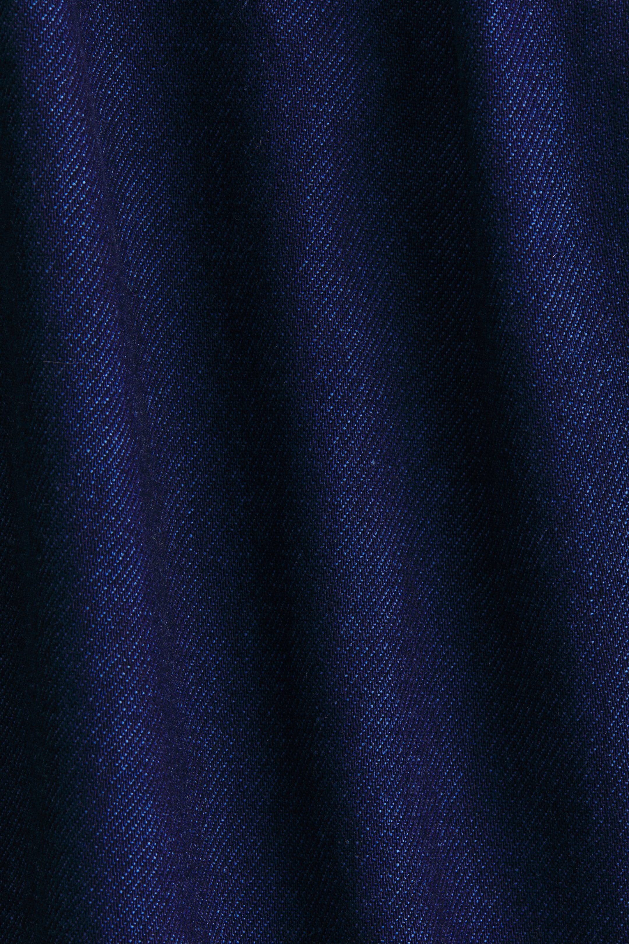 View larger image: A dark blue fabric is displaying a prominent diagonal twill weave pattern. Subtle folds in the material are creating variations, with lighter blue highlights contrasting against deeper navy shadows across the texture.