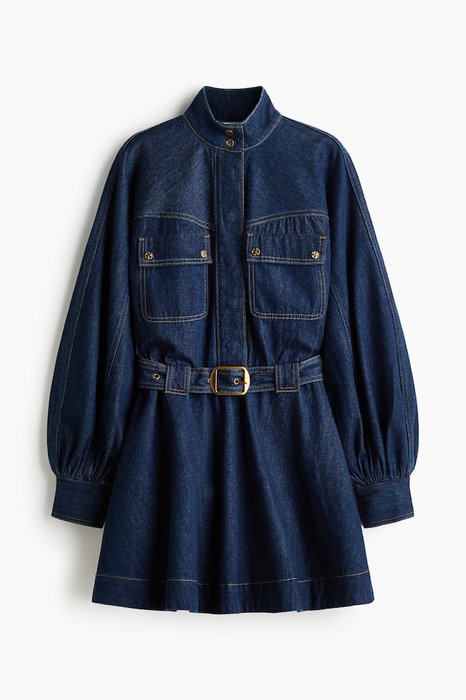 A dark blue denim dress is shown from the front, featuring a high stand collar, a concealed button placket, and two chest flap pockets with gold-toned studs. The dress features long balloon sleeves with wide cuffs, an adjustable waist belt with a gold-toned buckle, and a flared skirt. - 1