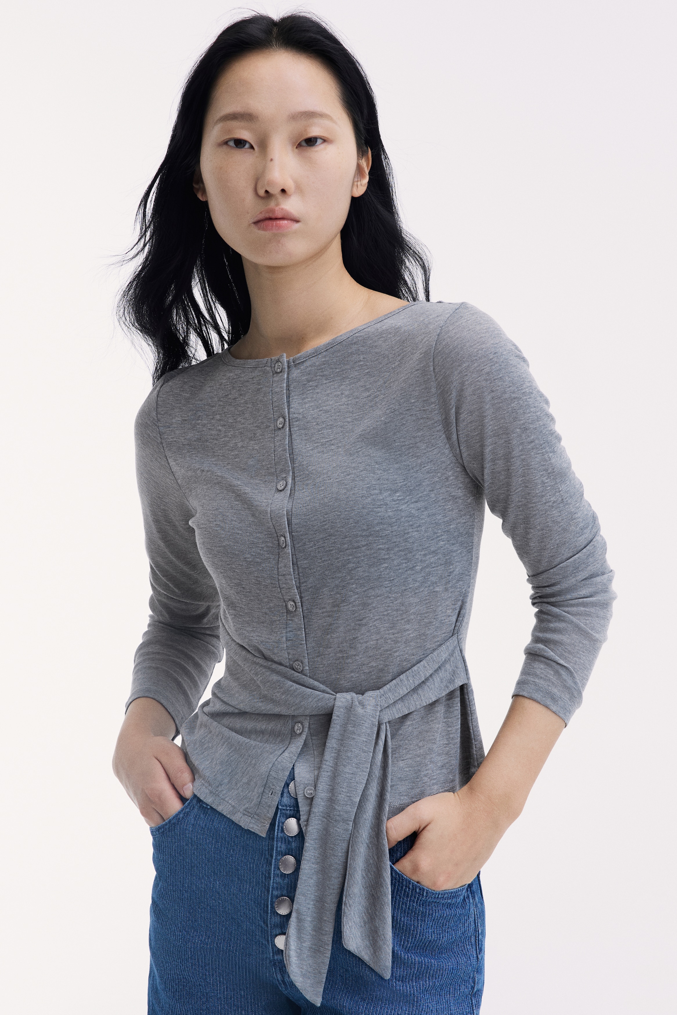 A heather grey, long-sleeved knit top is shown from the front, featuring a wide boat neckline and a button-down front closure. The top is styled with an asymmetrical fabric panel wrapping across the waist and fastening with a side tie.