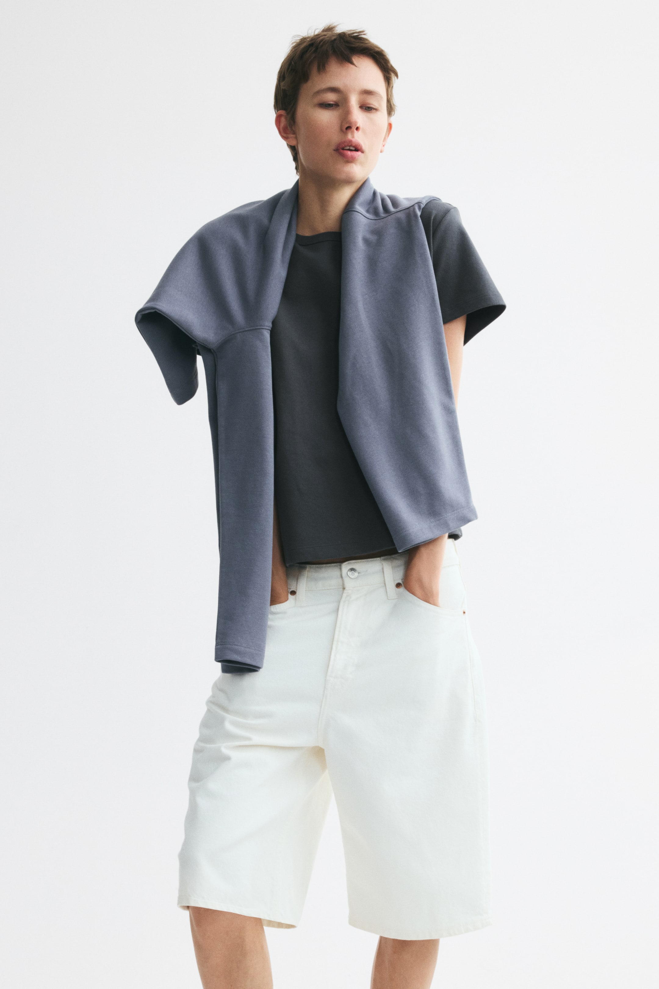 View larger image: A dark grey fitted T-shirt in heavyweight cotton jersey with ribbing around the neckline and a straight-cut hem is worn by the model. A blue-grey sweatshirt is draped over the model’s shoulders, partially covering the T-shirt.
