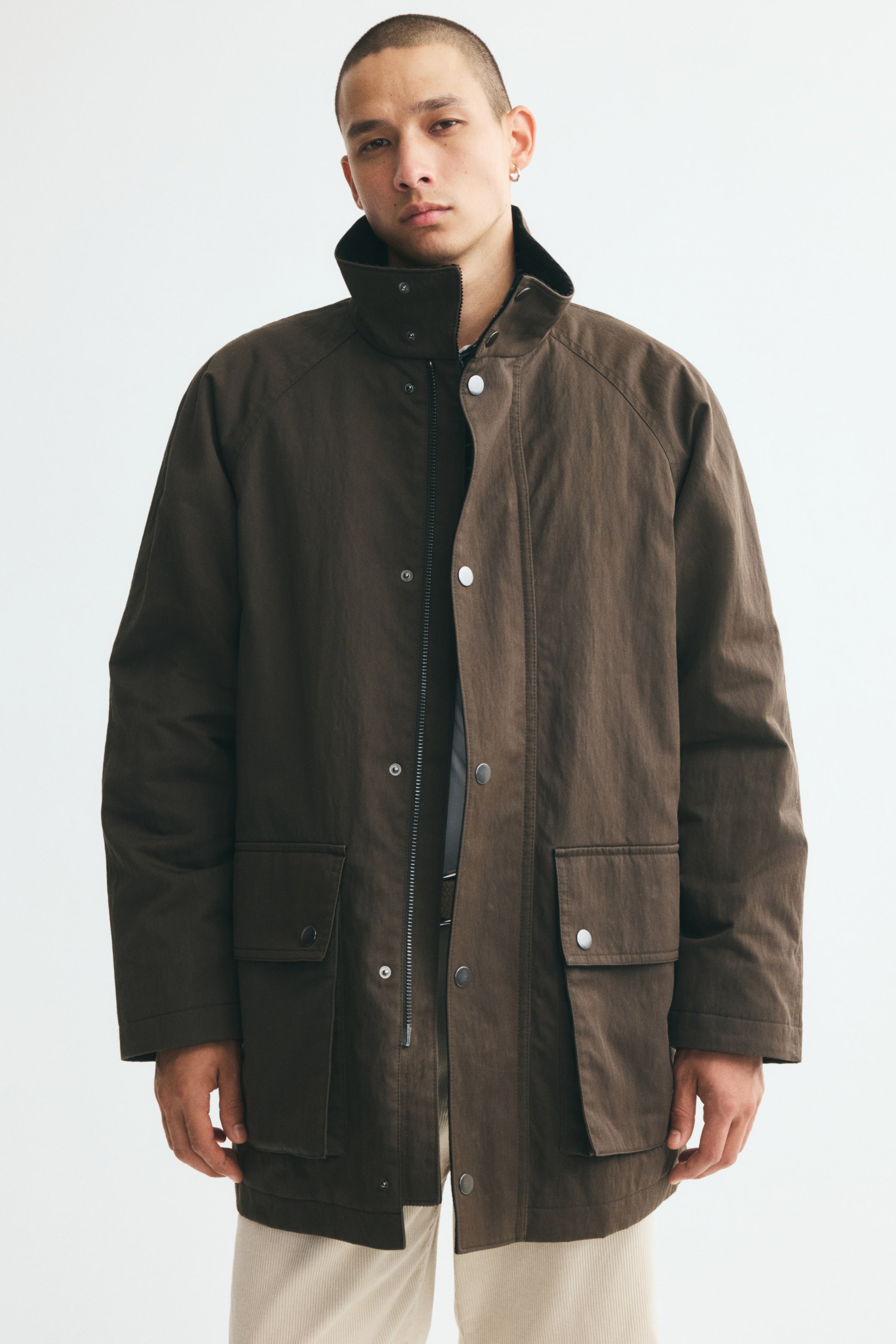 View larger image: The model is wearing a brown cotton-blend twill parka, featuring a raised stand collar and a partially unzipped front with a press-stud wind flap. The parka has long raglan sleeves and large flap front pockets, and is styled over a dark top and light-coloured trousers.