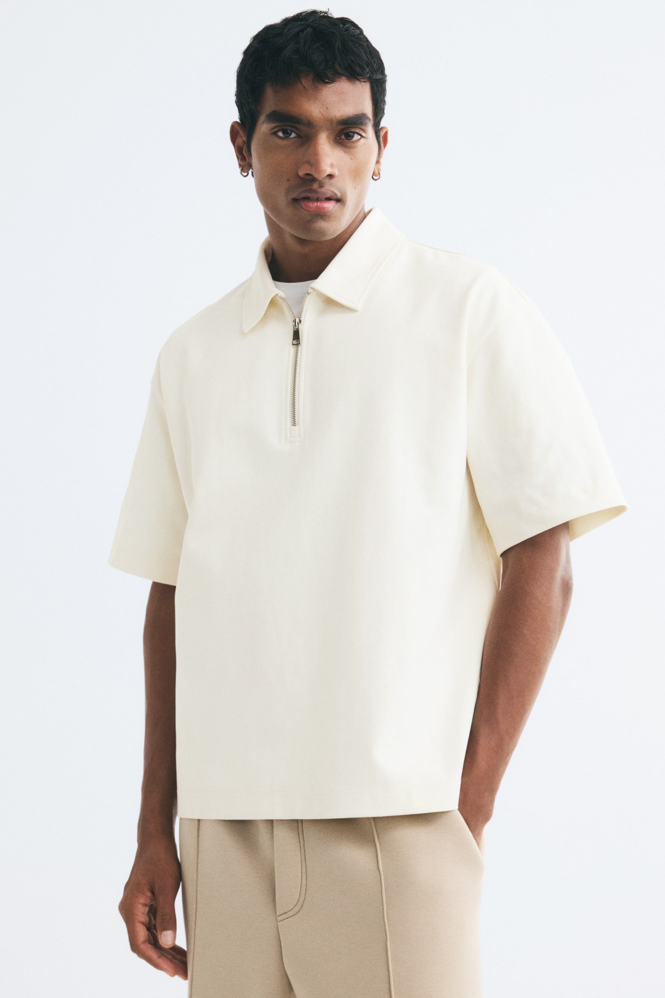 Xem ảnh lớn hơn: Một chiếc áo polo màu kem làm từ vải jersey cotton interlock đang được người mẫu mặc, với phom dáng thoải mái, cổ áo cổ điển, cổ kéo khóa nửa và tay áo ngắn rộng. Áo được phối layer bên ngoài một chiếc áo lót trắng và kết hợp với quần dài màu be.