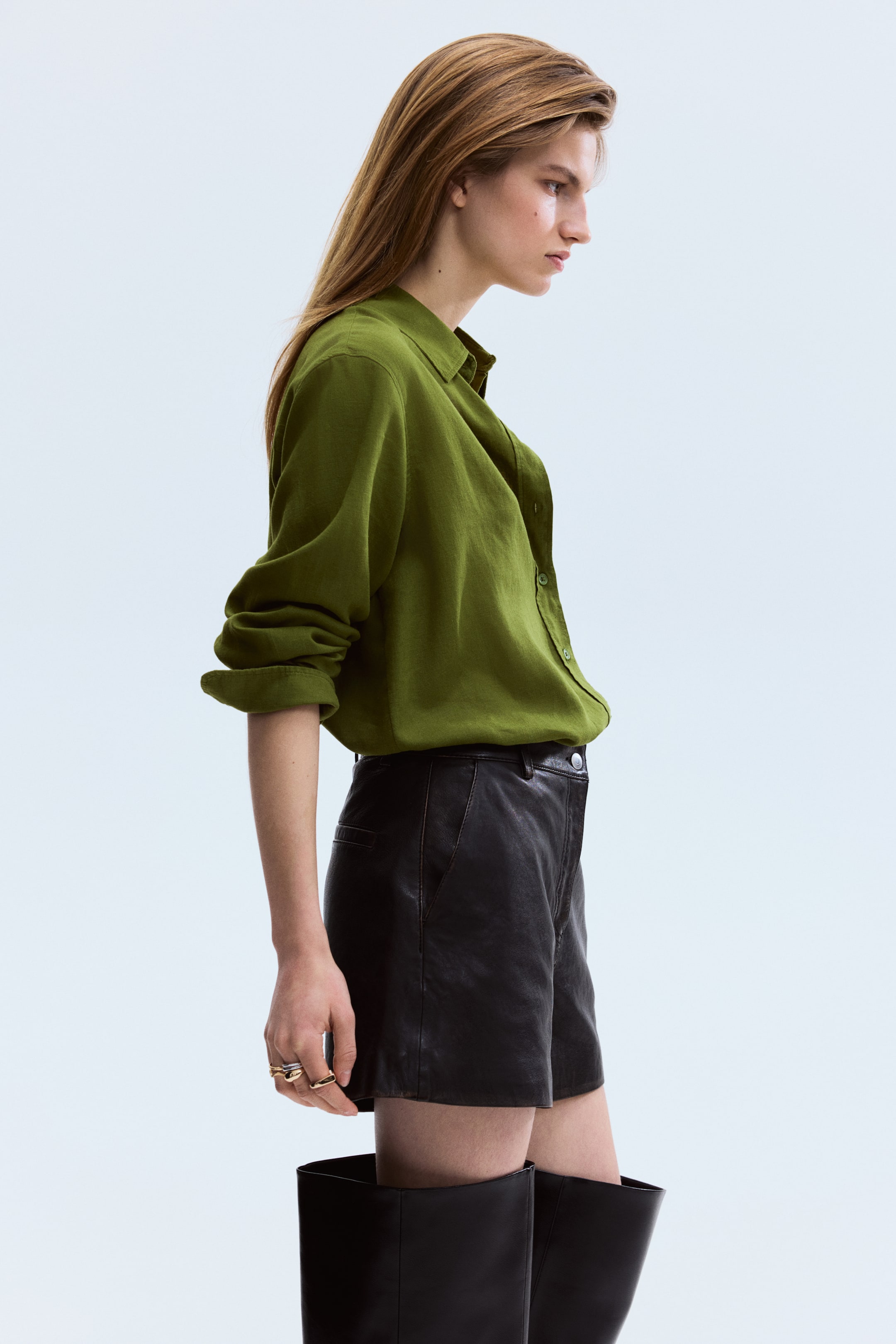 Agrandir l'image: Une chemise olive à boutons, confectionnée dans un tissu texturé, est présentée de biais, avec un col et des manches longues retroussées. Elle est partiellement rentrée dans un short noir ajusté, qui semble être en cuir avec une fermeture à boutons visible et des poches latérales.