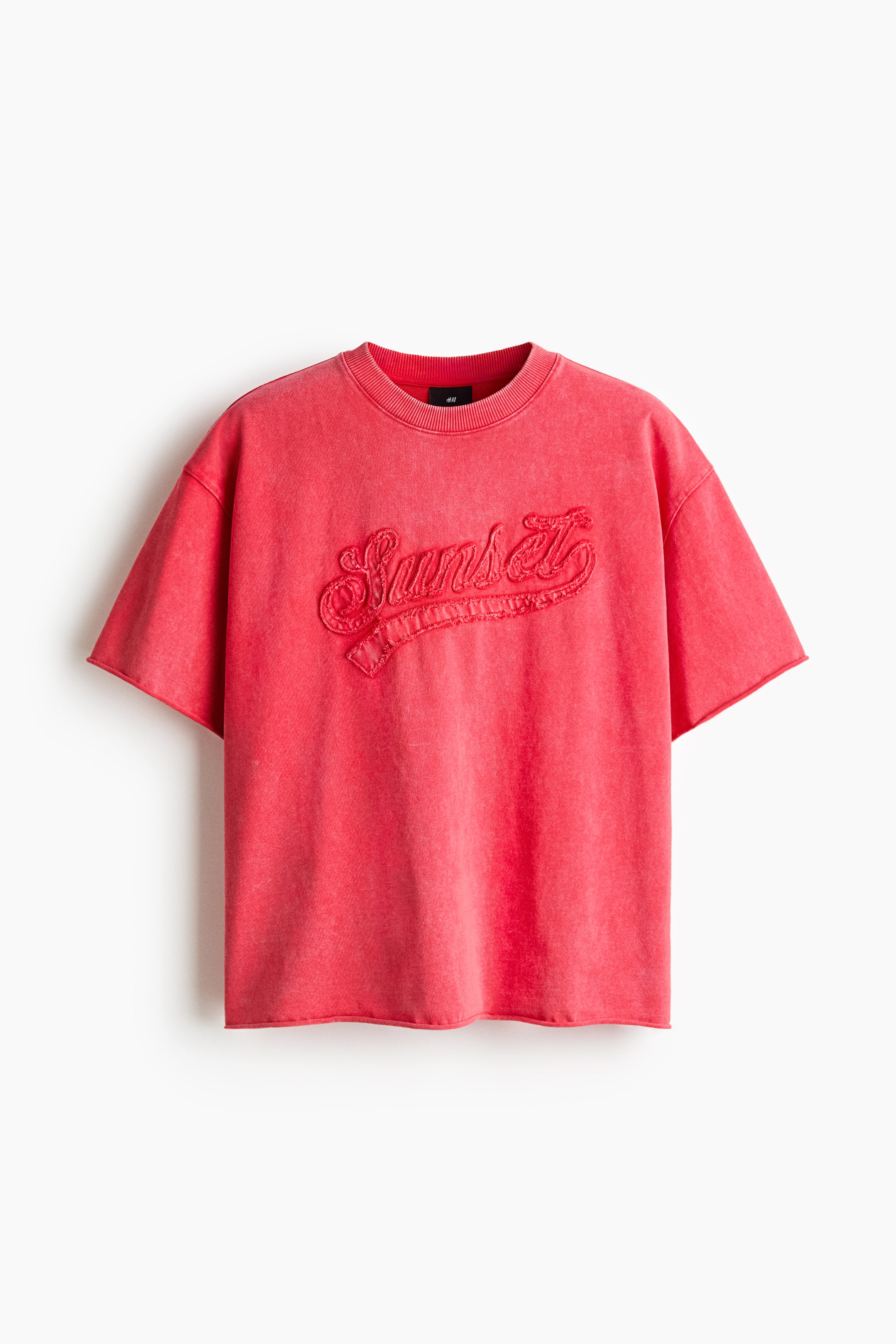 Se større billede: En T-shirt i bokset stil i vasket rød præsenterer sin front, der byder på en rund, ribkantet halsudskæring og nedsænkede skuldre. Fronten viser en broderet 'Sunset'-tekst, og de korte ærmer og den nederste kant har ubehandlede kanter.
