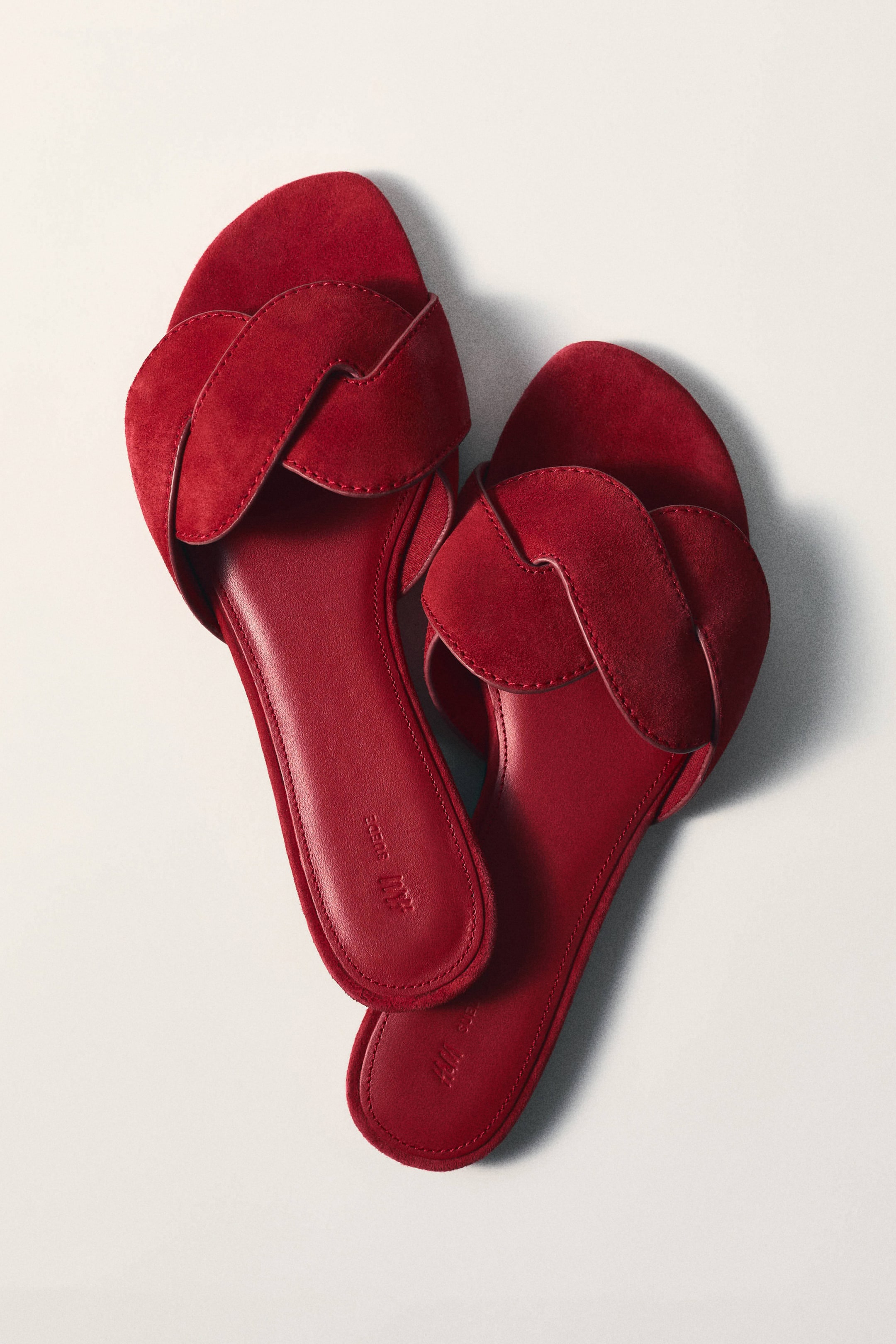 View larger image: Two red flat sandals are positioned at an angle, showcasing wide, overlapping suede straps across the instep. Each sandal features a flat footbed with visible stitching and the embossed text 'H&M Suede'.