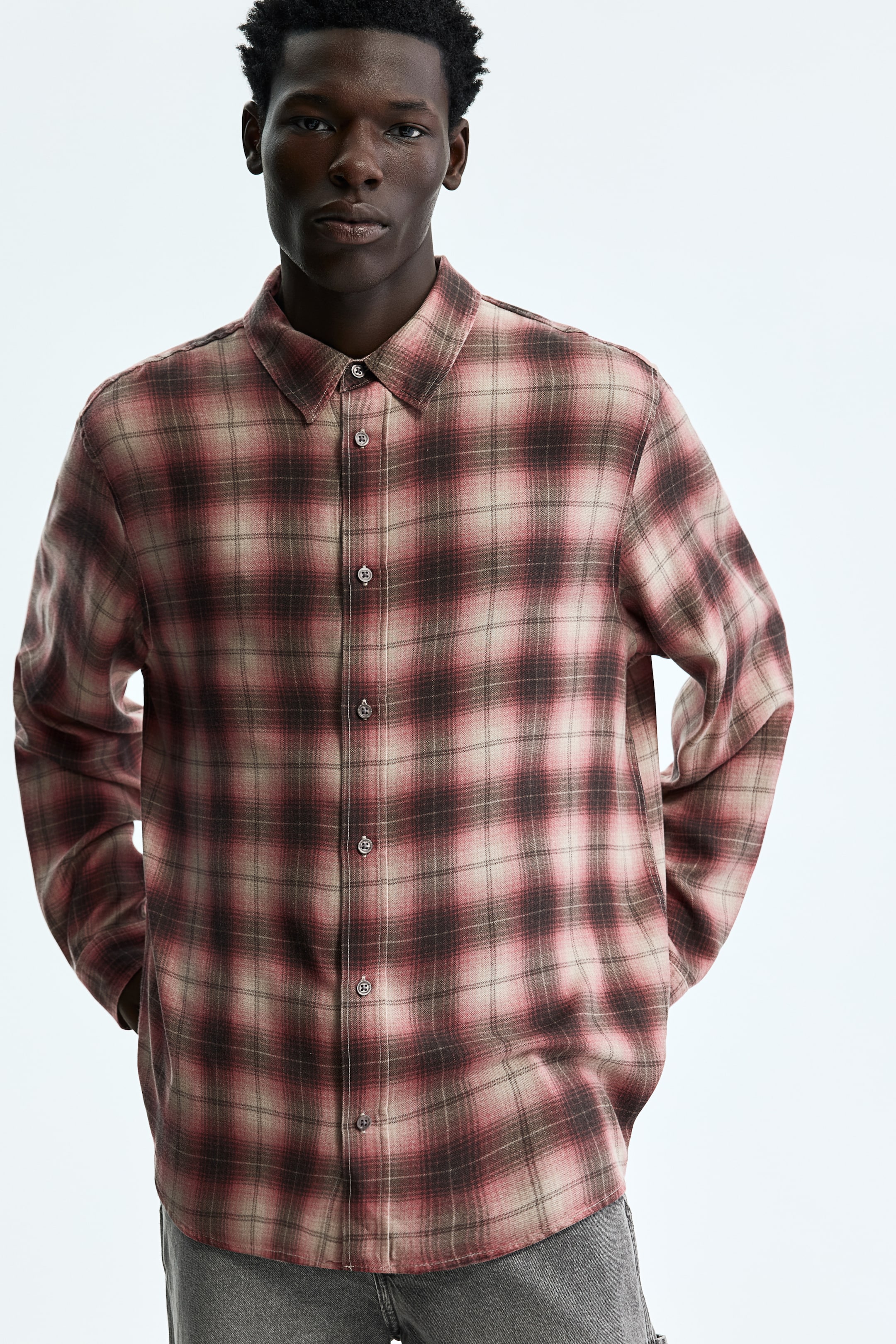 View larger image: A red, brown, and beige plaid button-up shirt is worn, featuring a pointed collar and long sleeves. The shirt showcases a relaxed fit, with its rounded hem falling just below the waist.