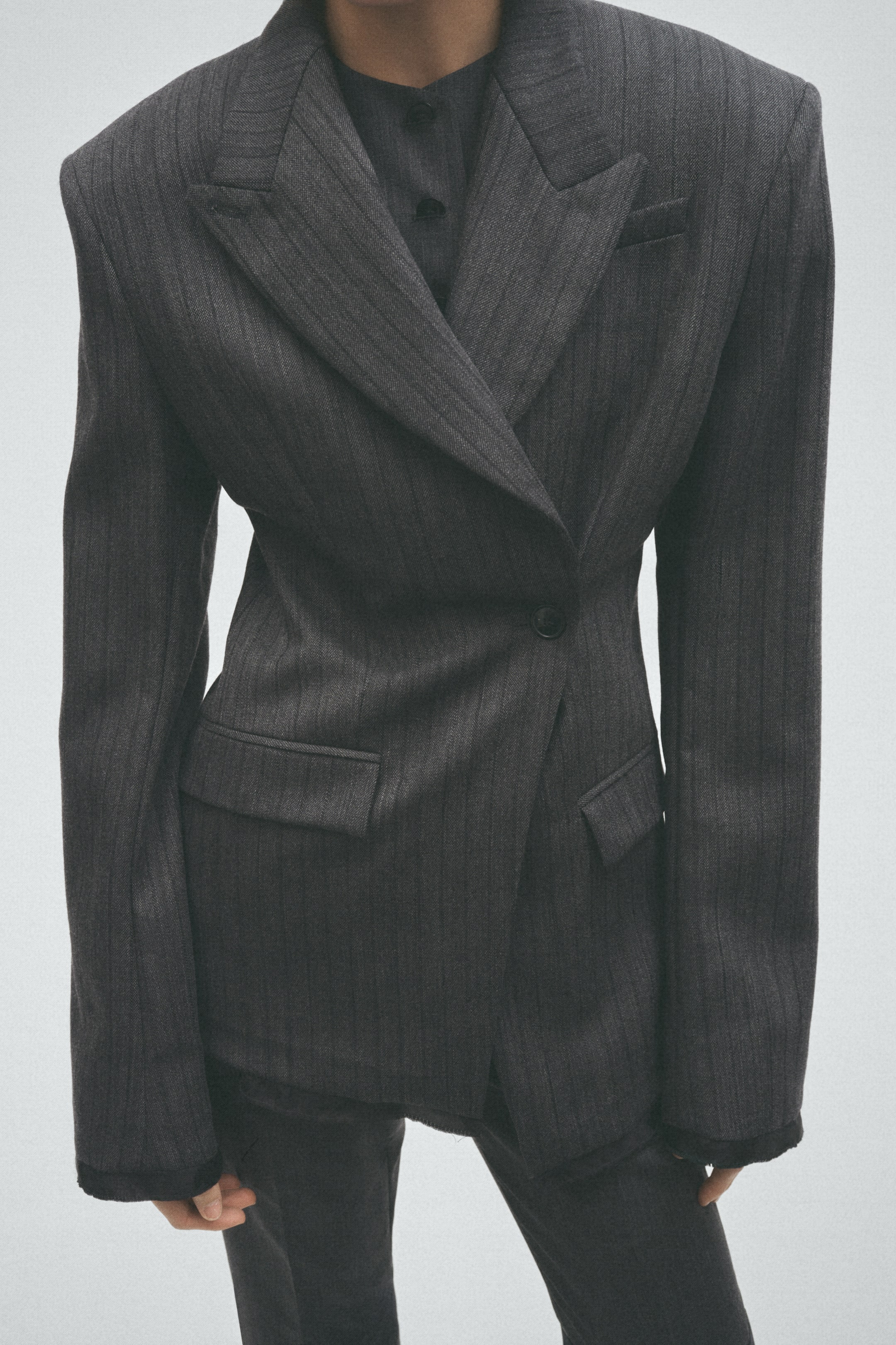 View larger image: The model is wearing a dark grey pinstripe blazer. It features a double-breasted front with a single-button closure, structured shoulders, notched lapels, and flap pockets, layered over a dark button-front shirt.
