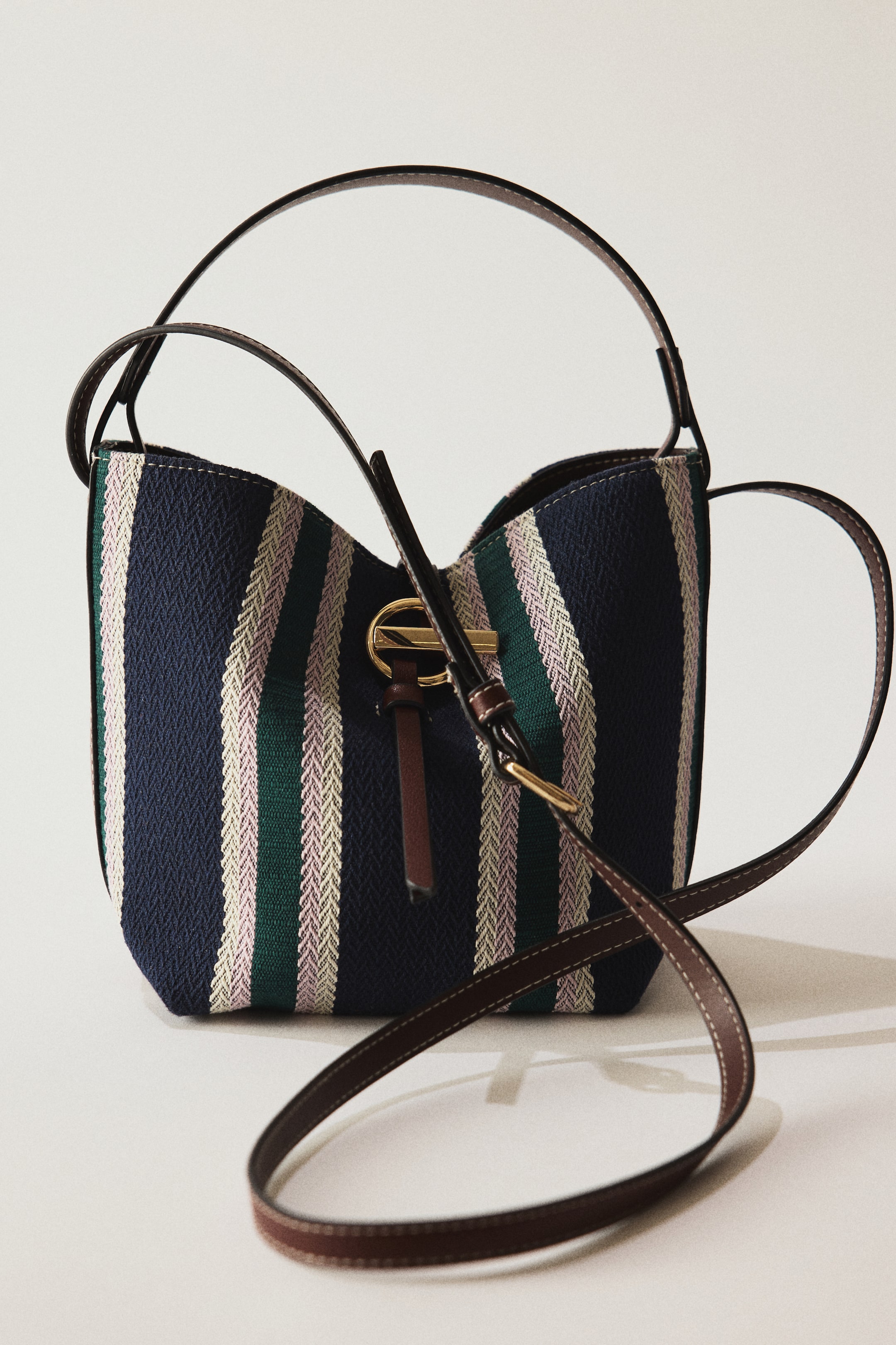 Grösseres Bild ansehen: Eine Bucket-Bag steht, leicht nach rechts geneigt, und zeigt vertikale, gewebte Streifen in Navyblau, Dunkelgrün und zartrosa. Sie verfügt über schlanke, dunkelbraune Lederhenkel und einen langen Crossbody-Riemen; auf der Vorderseite sind ein goldfarbener Ring sowie ein kurzes braunes Lederdetail sichtbar.