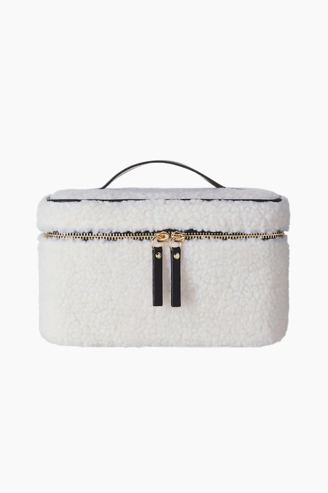 Une trousse de maquillage de forme carrÃ©e en matiÃ¨re teddy texturÃ©e Ã©cru est prÃ©sentÃ©e de face, mettant en valeur une fermeture zippÃ©e ton or avec deux tirettes en similicuir noir. Une fine poignÃ©e en similicuir noir arquÃ© sur le dessus du couvercle. - 1