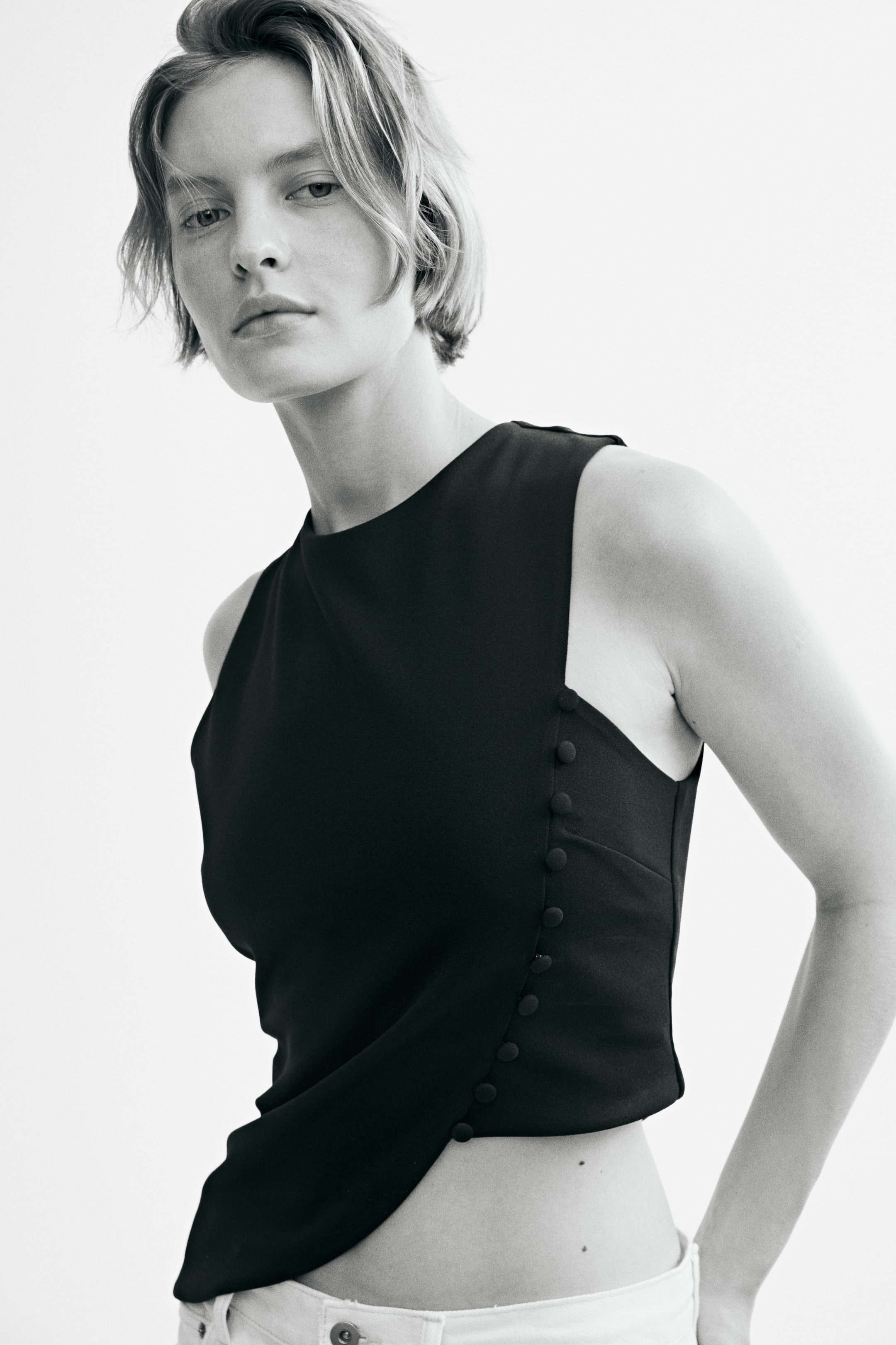 View larger image: The model is wearing a sleeveless top with a round neckline and an asymmetrical hem. A visible seam with decorative covered buttons runs along one side of the front, and the top is styled with light-coloured pants.