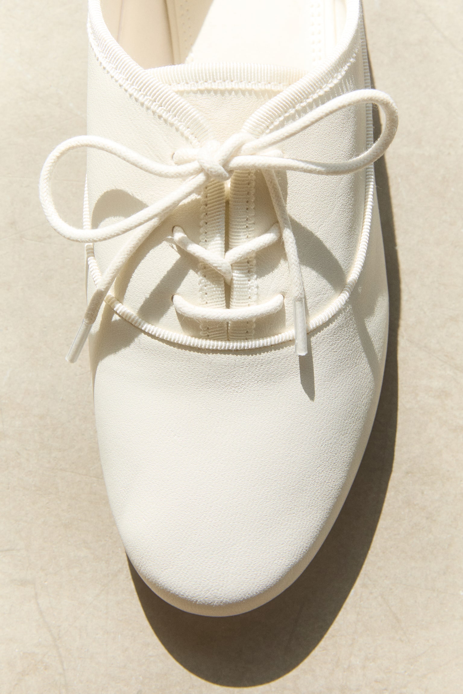 Off-white lace-up flats are showcased from a top-down angle, facing upwards. Each shoe boasts a sleek upper with a rounded toe, a tie-front lacing system with slender laces tied in a bow, a ribbed elasticated trim around the collar, and a small fabric pull tab at the heel. - 1