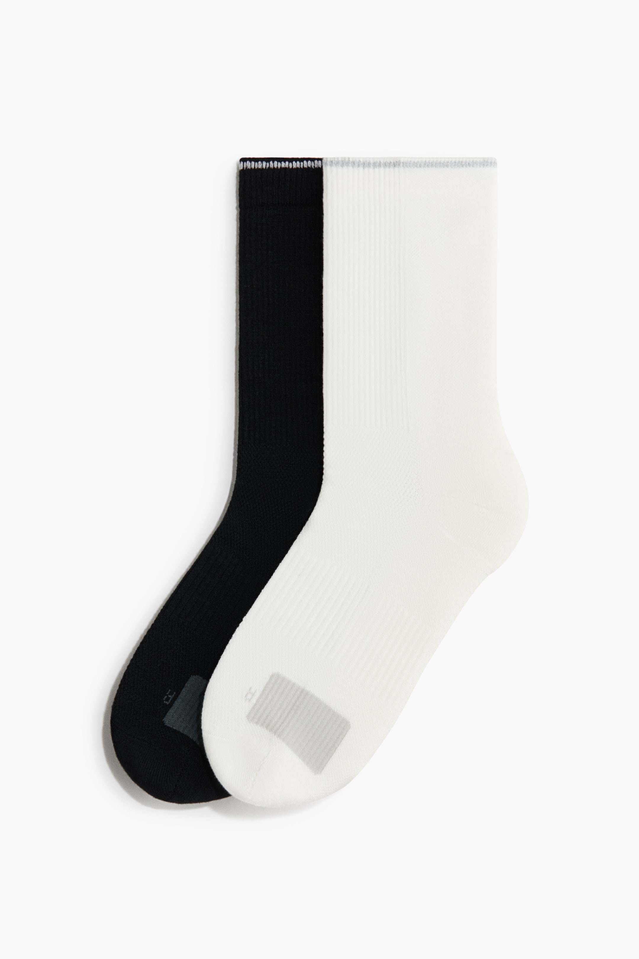 View larger image: Two crew-length socks, one black and one white, are laid flat in a side-profile view, with the white sock overlapping the black one. Both socks feature a ribbed leg texture and a contrasting grey rectangular knit patch on the arch.
