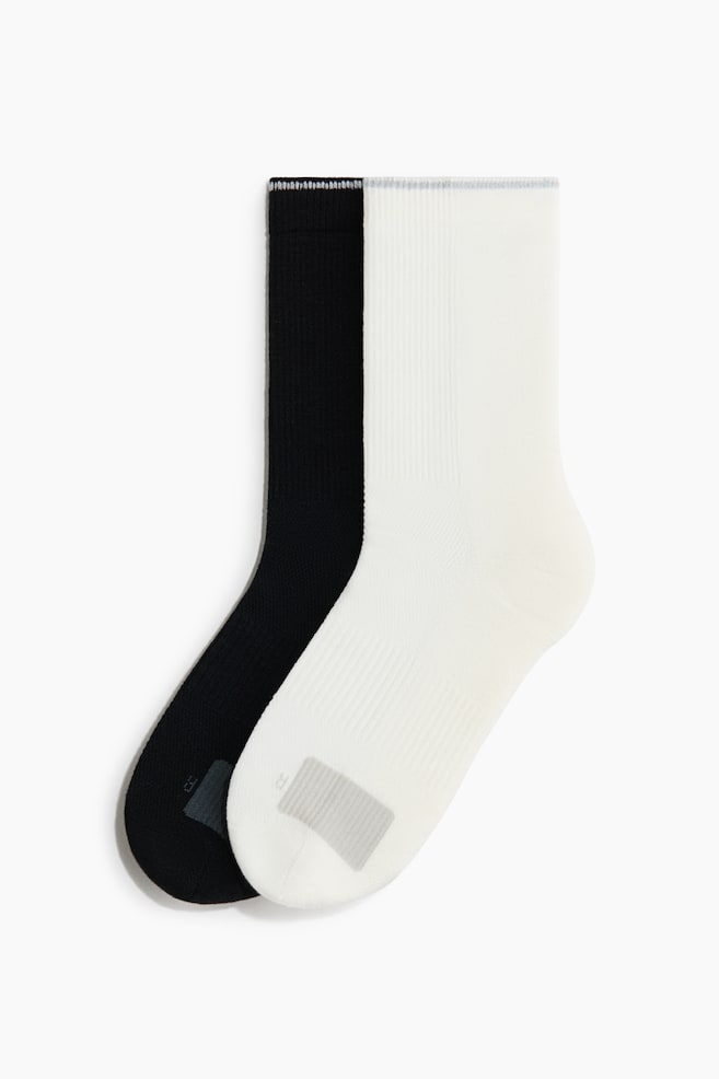 Two crew-length socks, one black and one white, are laid flat in side profile, with the white sock overlapping the black one. Both socks feature a ribbed leg texture and a contrasting grey rectangular knit patch on the arch. - 1