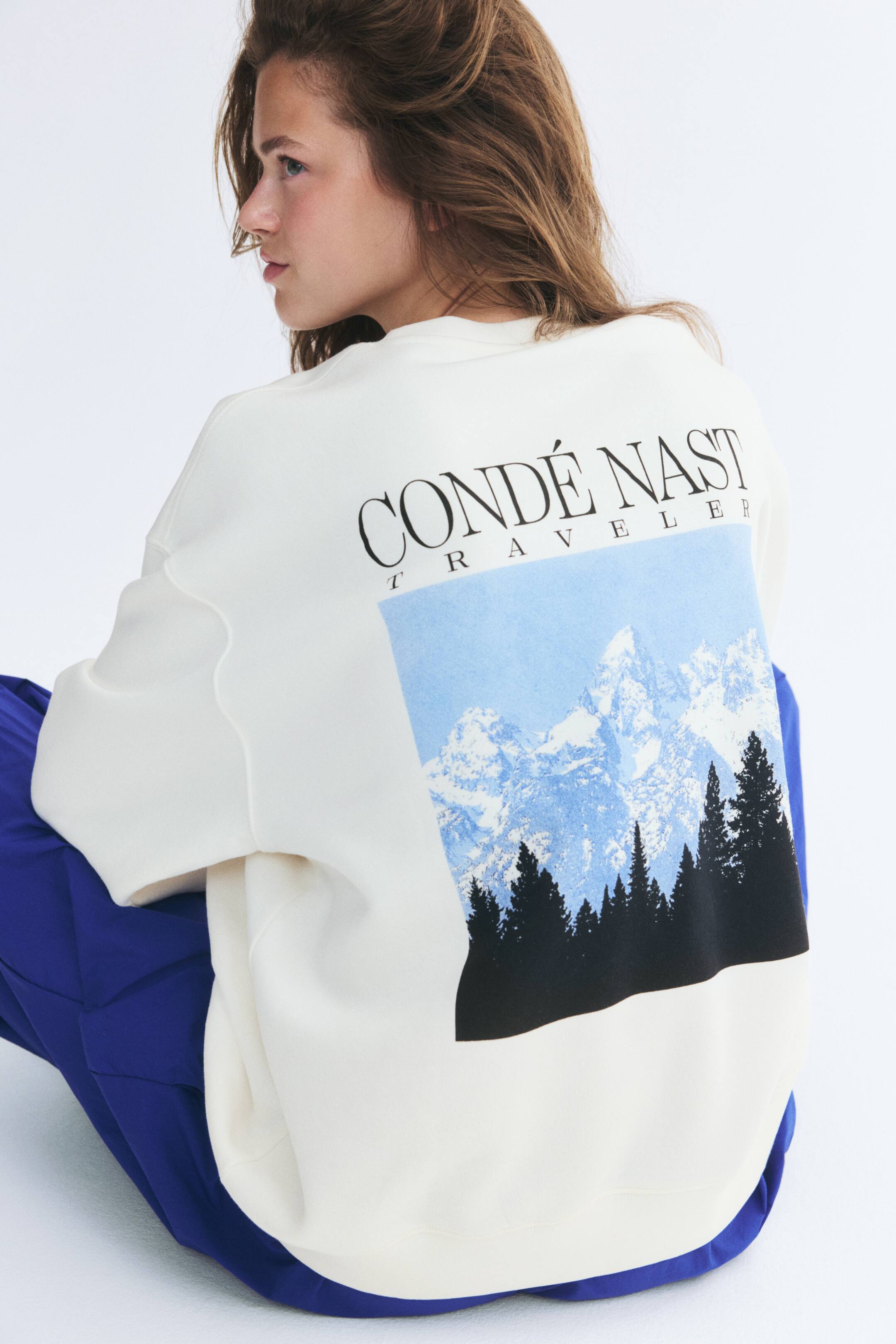 Xem ảnh lớn hơn: Một chiếc áo len fleece màu trắng ngà form oversized với cổ tròn, vai trễ và họa tiết in lớn ở mặt sau đang được người mẫu mặc. Họa tiết mô tả phong cảnh núi màu xanh với những cây thông bên dưới dòng chữ "CONDÉ NAST TRAVELER", và chiếc áo len này được phối cùng quần xanh lam.