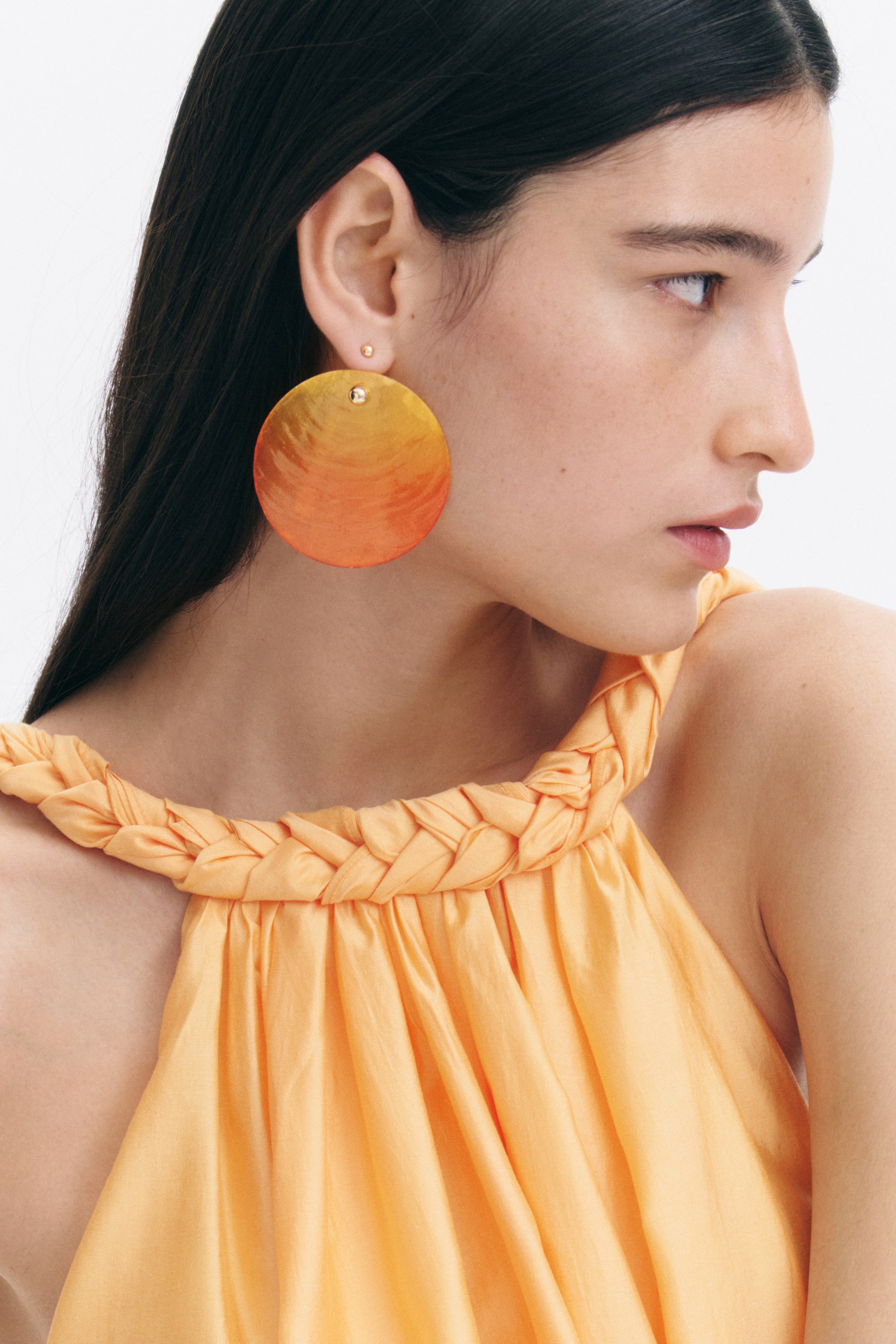 View larger image: The model is wearing statement earrings, each showcasing a large, circular, slightly translucent seashell pendant. The pendant features an orange and yellow ombré finish with a ridged texture and is secured to the earlobe by a small gold stud.