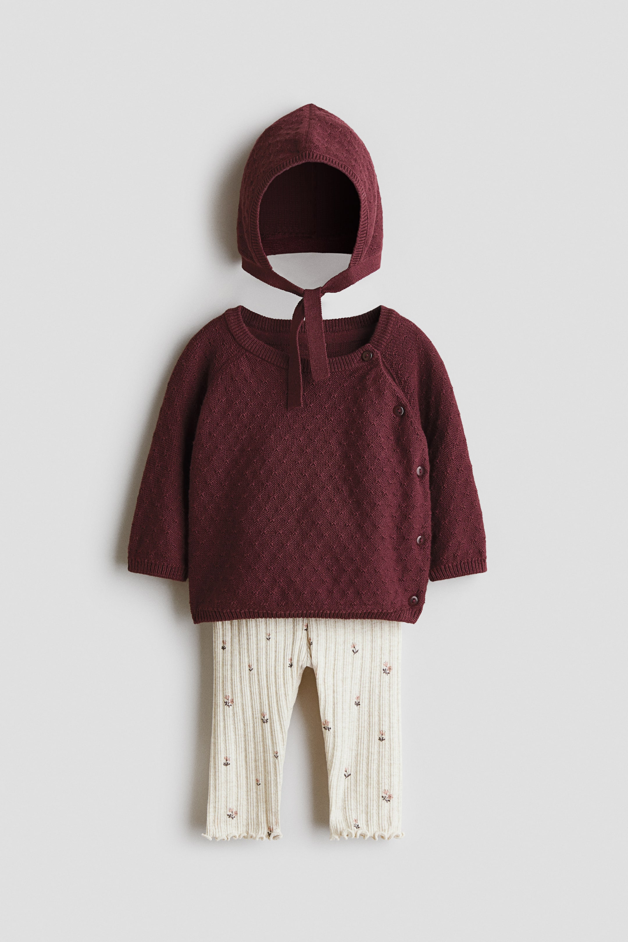 View larger image: A knitted baby outfit features a maroon bonnet with ties and a long-sleeved sweater with a textured pattern and asymmetrical button closure on the right side. Coordinating cream ribbed leggings with small floral embroidery and ruffled hems complement the sweater.