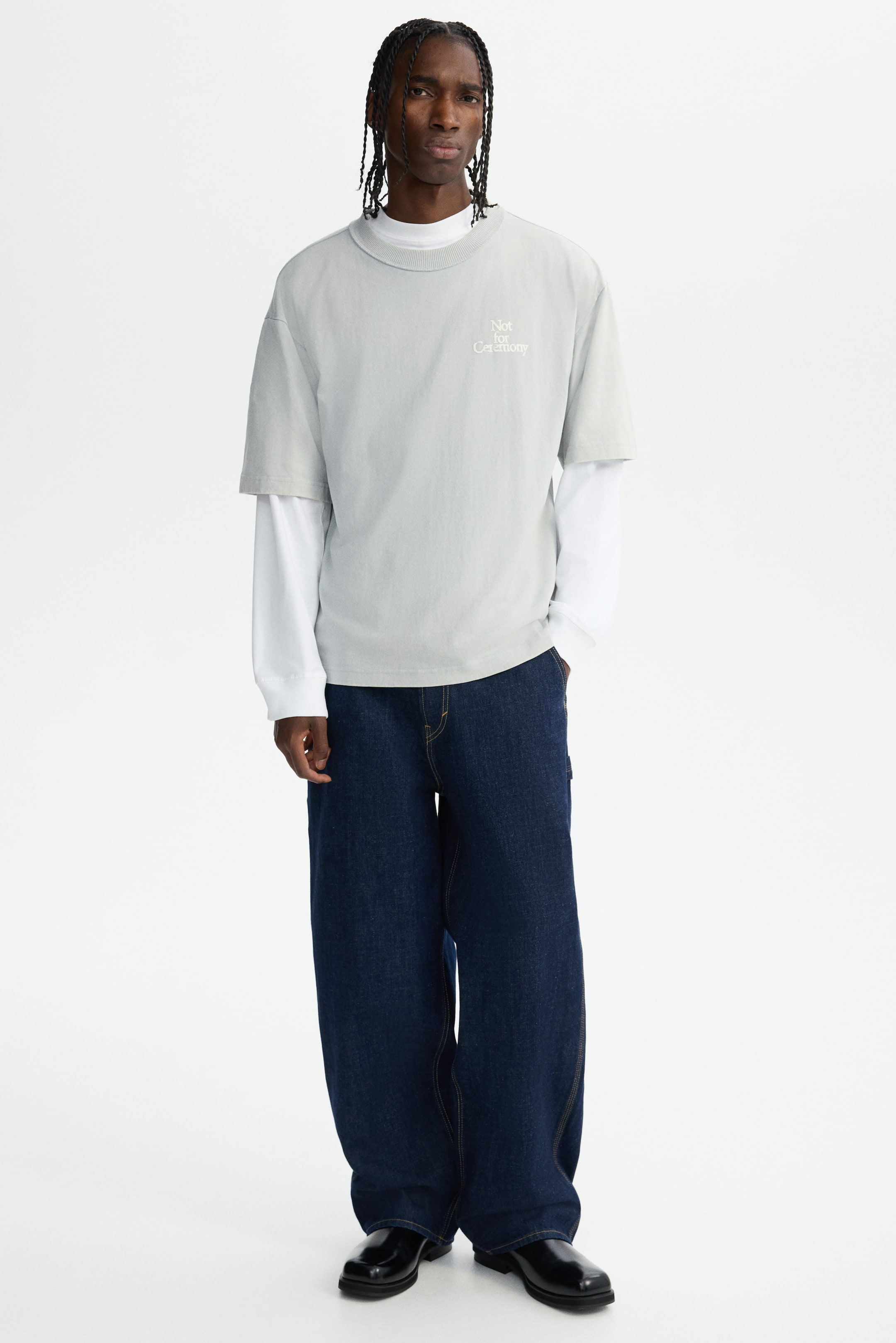 View larger image: The product, a light gray oversized short-sleeve t-shirt with a ribbed crew neck and 'Not for Ceremony' text on the chest, is being layered over a white long-sleeve shirt. Dark blue wide-leg jeans with contrasting yellow stitching are also being worn, all presented from a full front view.